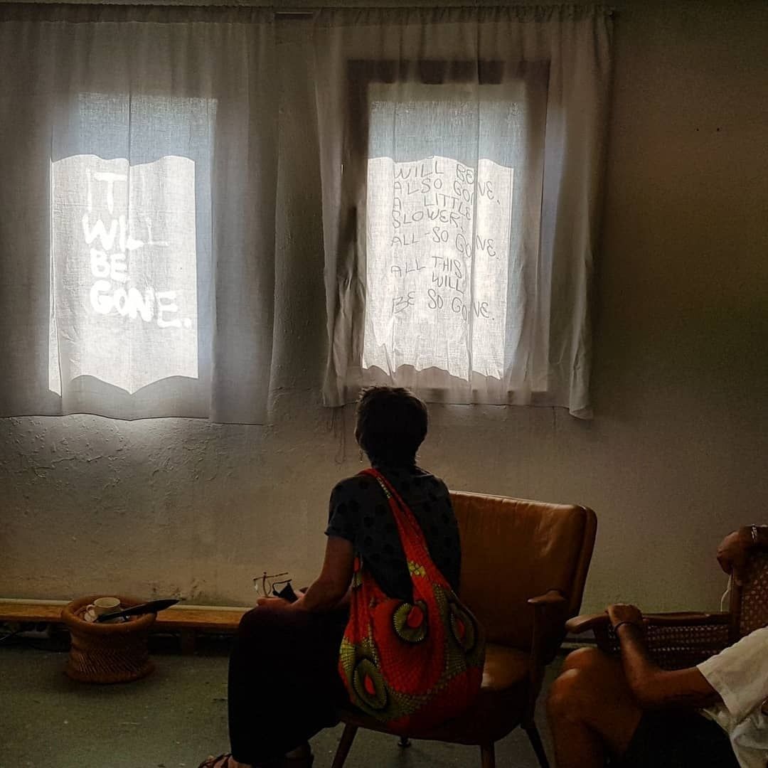 A person seen from behind in a brown leather armchair, a red patterned bag over the shoulder, looking at the two curtained windows with writing lit from behind; at the right edge the arm and knee of a second person.