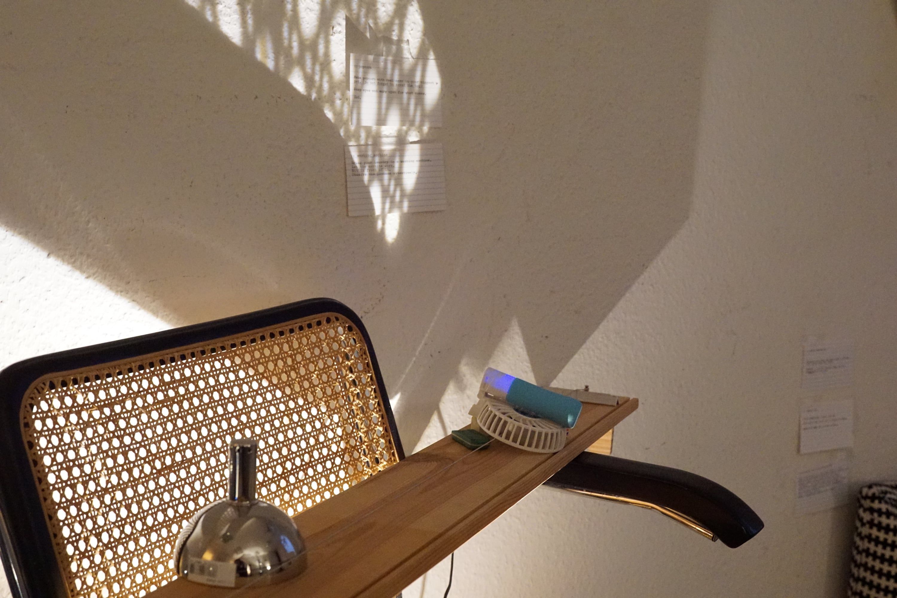 The cane back of a chair in warm light, on its arm a wooden board with a small fan and a chrome bell, index cards with typed text on the wall behind
