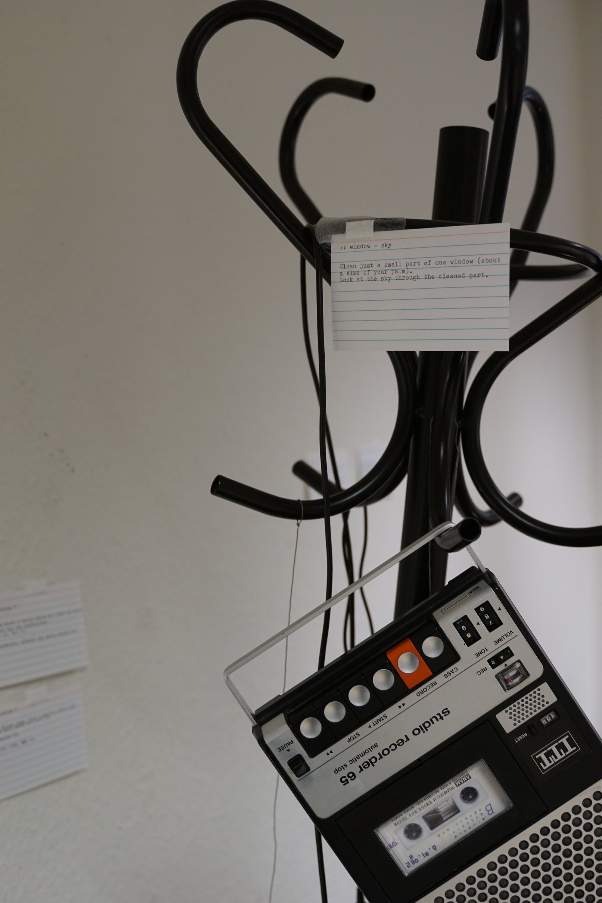 A black bentwood coat stand against a white wall, an index card with typed text on one hook and an old cassette recorder hanging at the bottom
