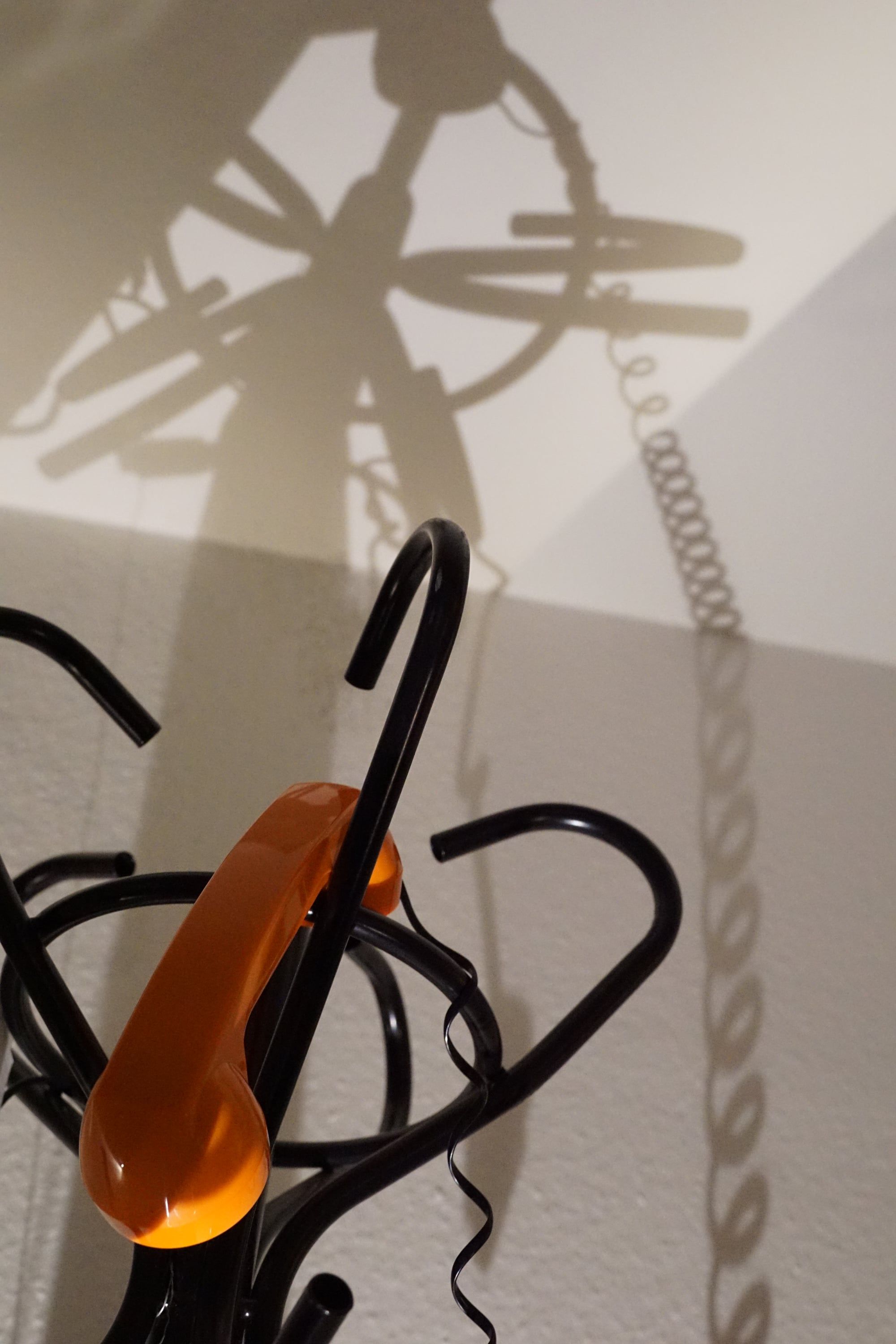The hooks of a black coat stand with an orange telephone handset hanging from one of them, the long shadow of the stand and a coiled cord on the wall behind