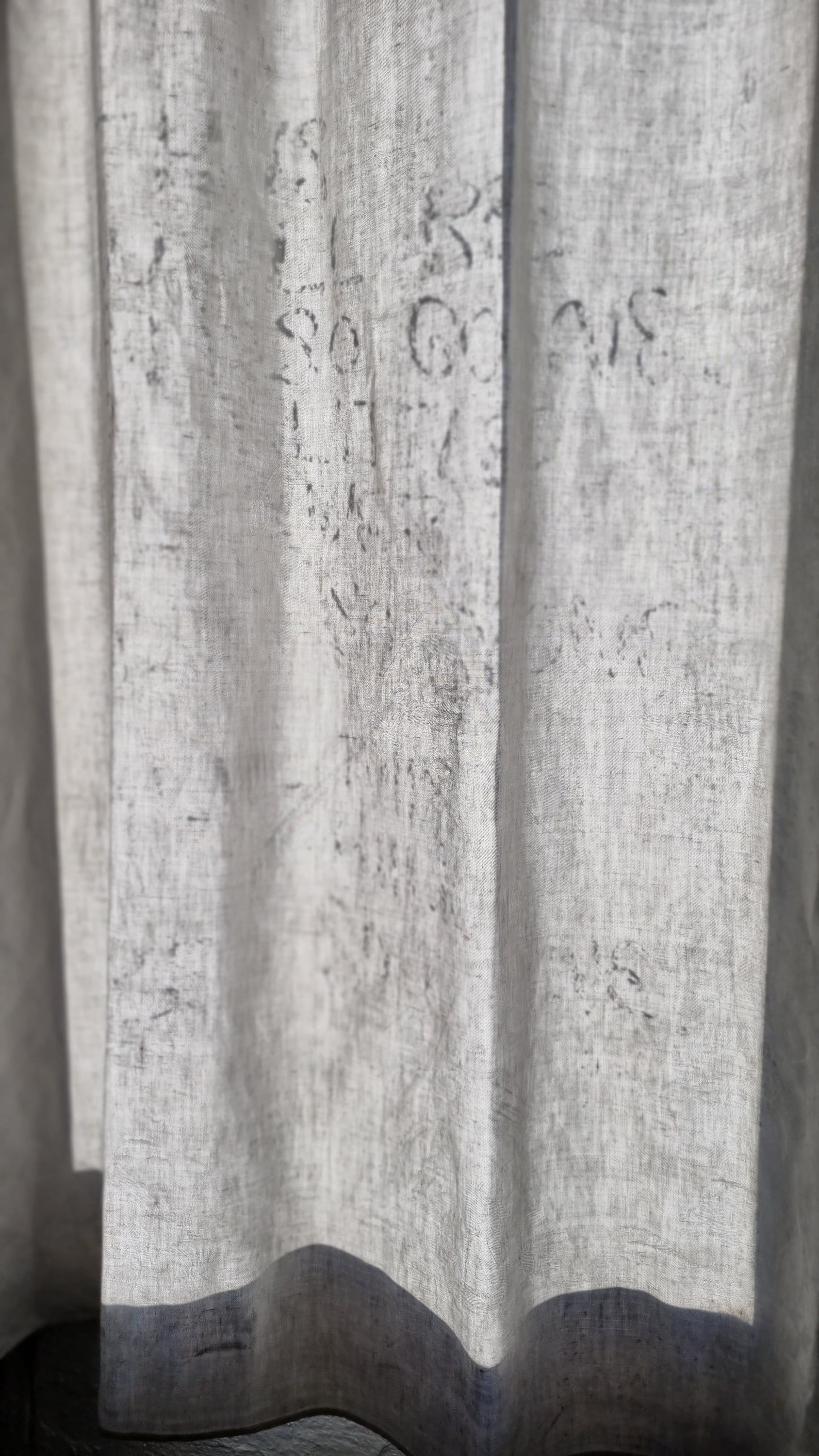 A tall sheer curtain in sunlight with faint grey traces of writing, its hem casting a wavy shadow.
