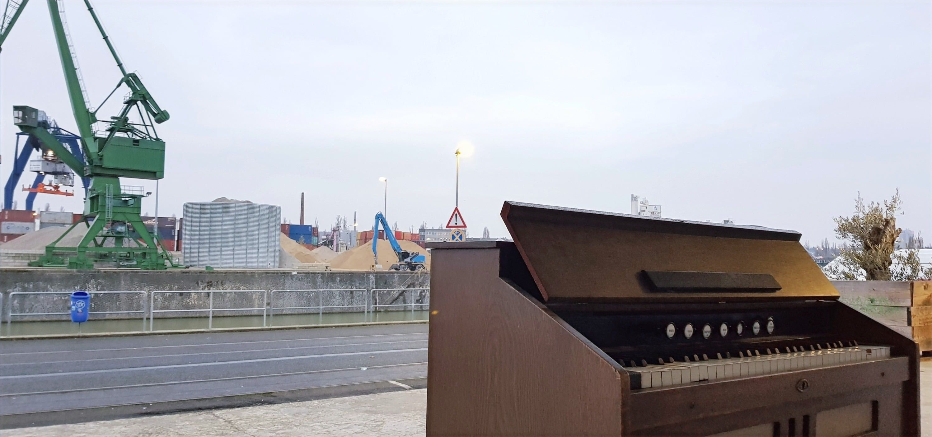 Ein braunes Harmonium am Hafen, dahinter ein grüner Kran und ein bewölkter Himmel.