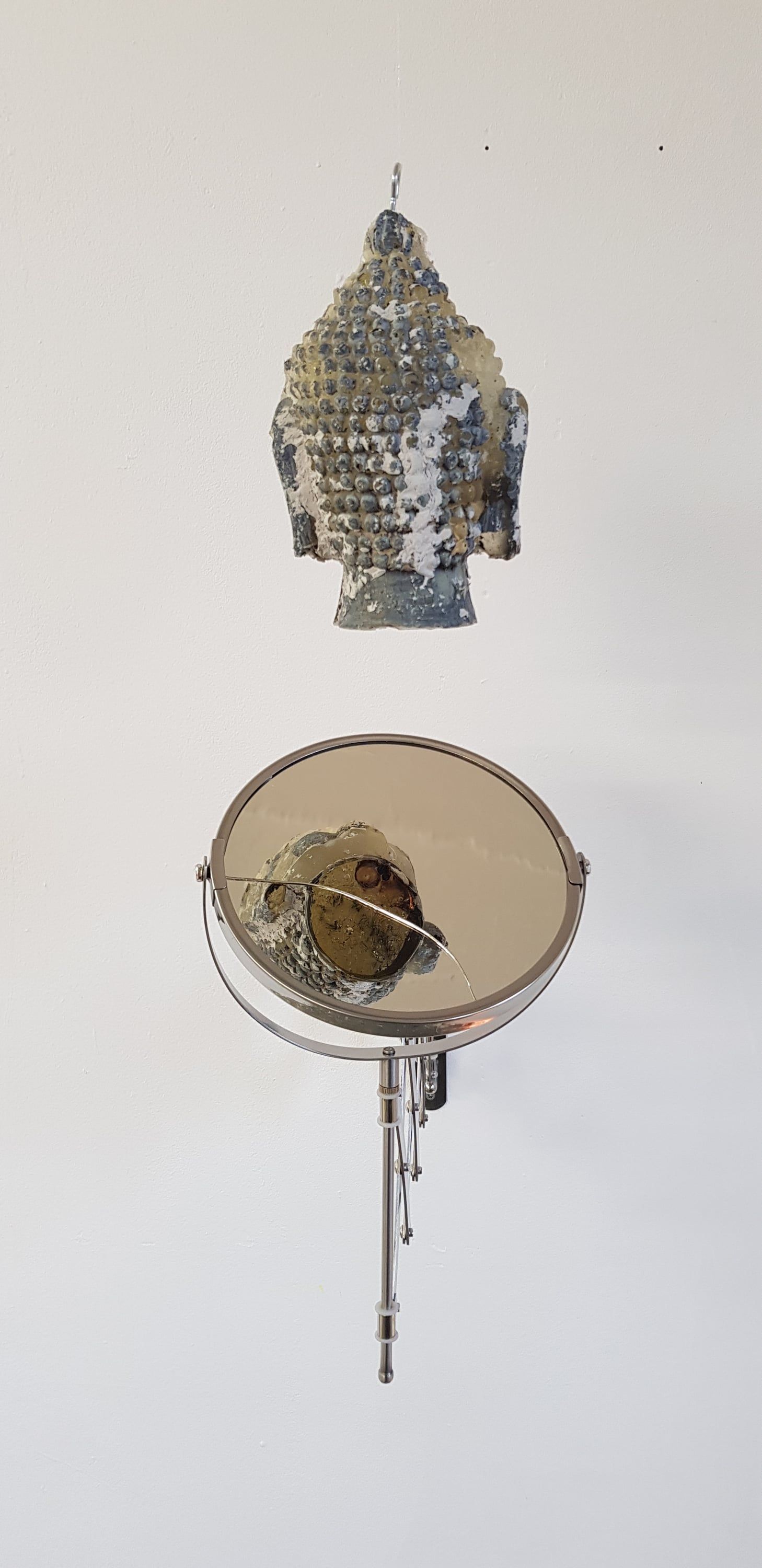 The resin head hangs high on a white wall; below it a round mirror on a thin stand shows the head and a crack of light across its glass.