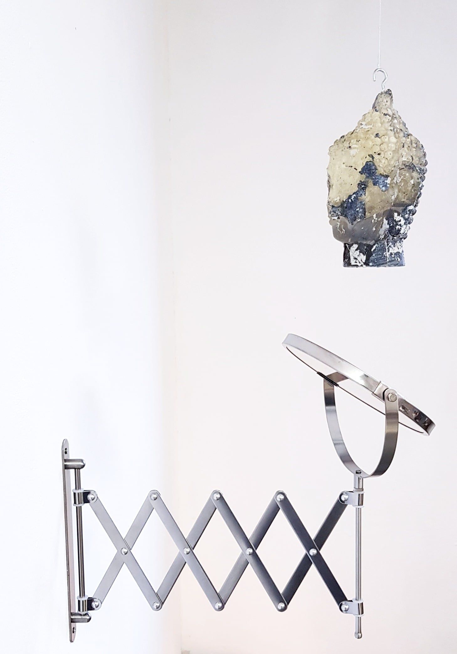 A head with a Buddha's curled hair, cast in translucent resin with blue-grey and white patches, hangs from a hook above a round steel mirror on an extendable scissor arm, against a white wall.