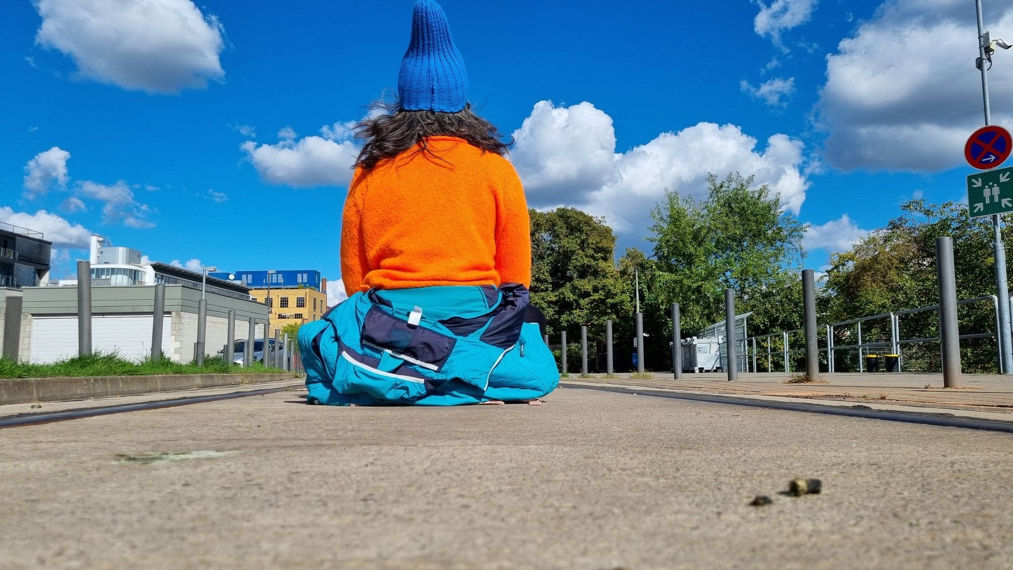 A person in an orange sweater and a blue woolly hat sits cross-legged on a paved square, seen from behind, under a blue sky with clouds; two small bird droppings lie on the ground in front of them.