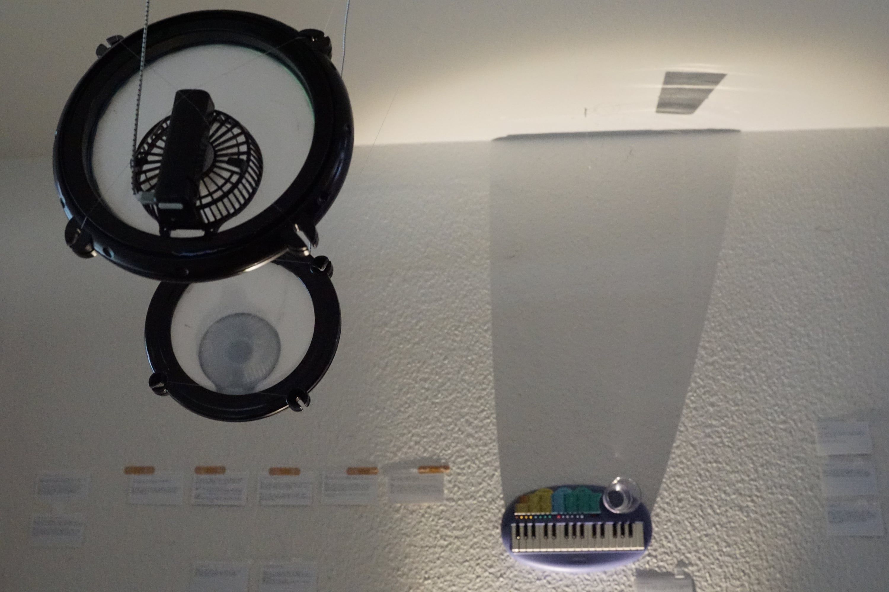 Two black hoops with small fans hanging from the ceiling, below them on the wall a pale band of light that ends at a small keyboard, index cards along the wall