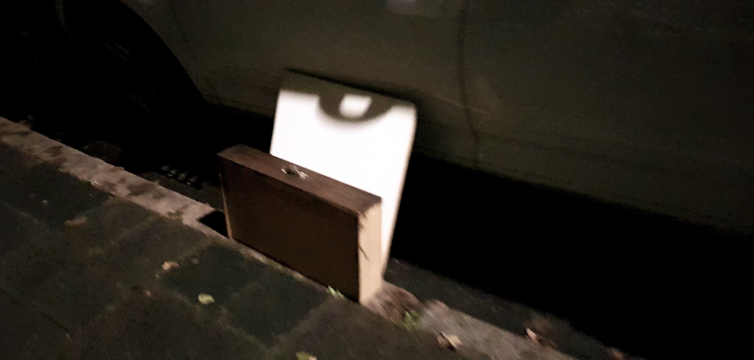 Ein brauner Koffer lehnt im Dunkeln an einer Wand, über ihm ein heller Fleck.