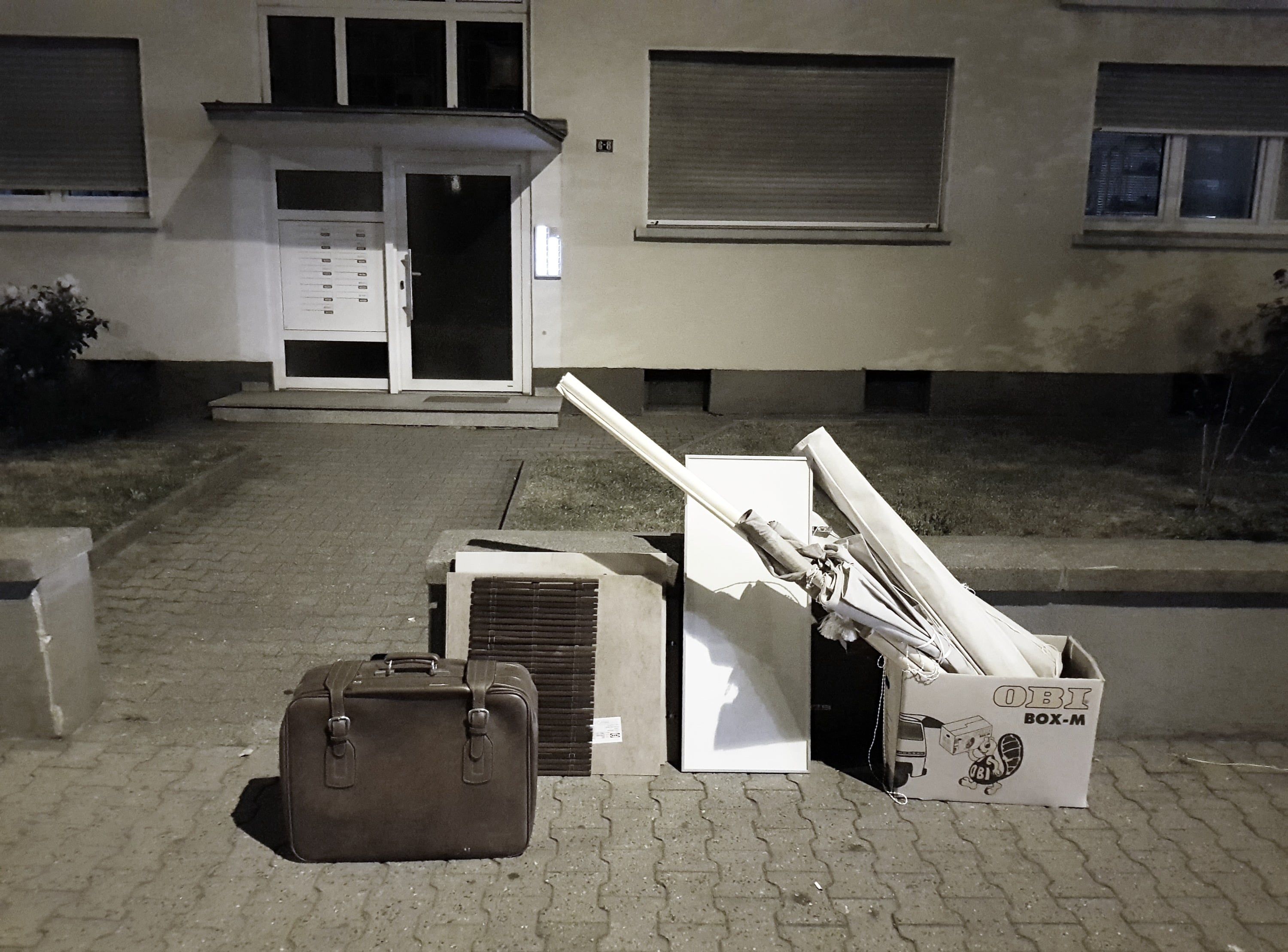 Abends auf dem Gehweg vor einem Wohnhaus: ein dunkelbrauner Koffer neben weiteren abgestellten Dingen, darunter ein weißer Schrank und ein offener Karton mit Stangen.