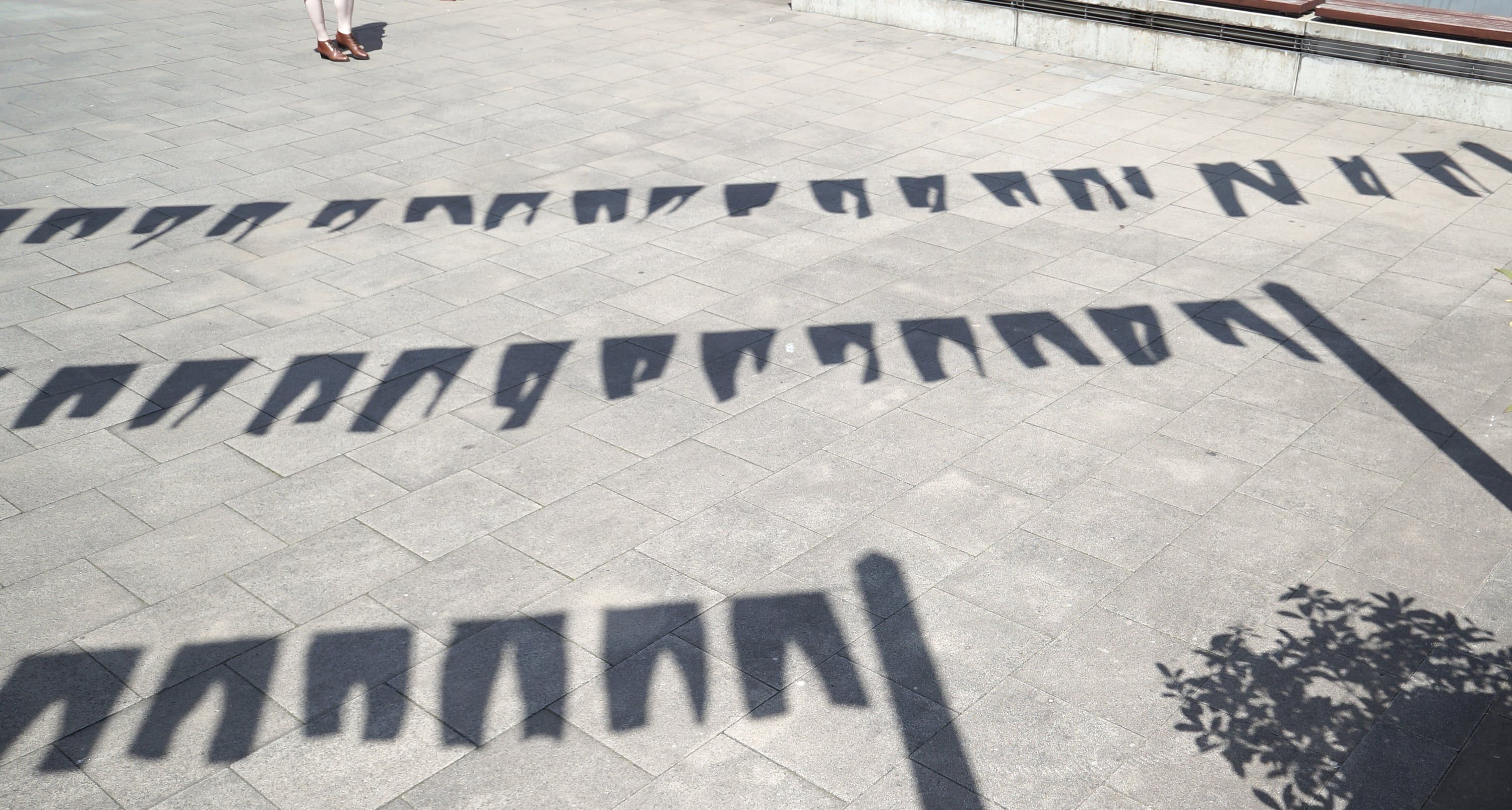 Drei Reihen von Schatten der aufgehängten Leggings auf grauem Pflaster; am oberen Bildrand stehen die Beine einer Person.
