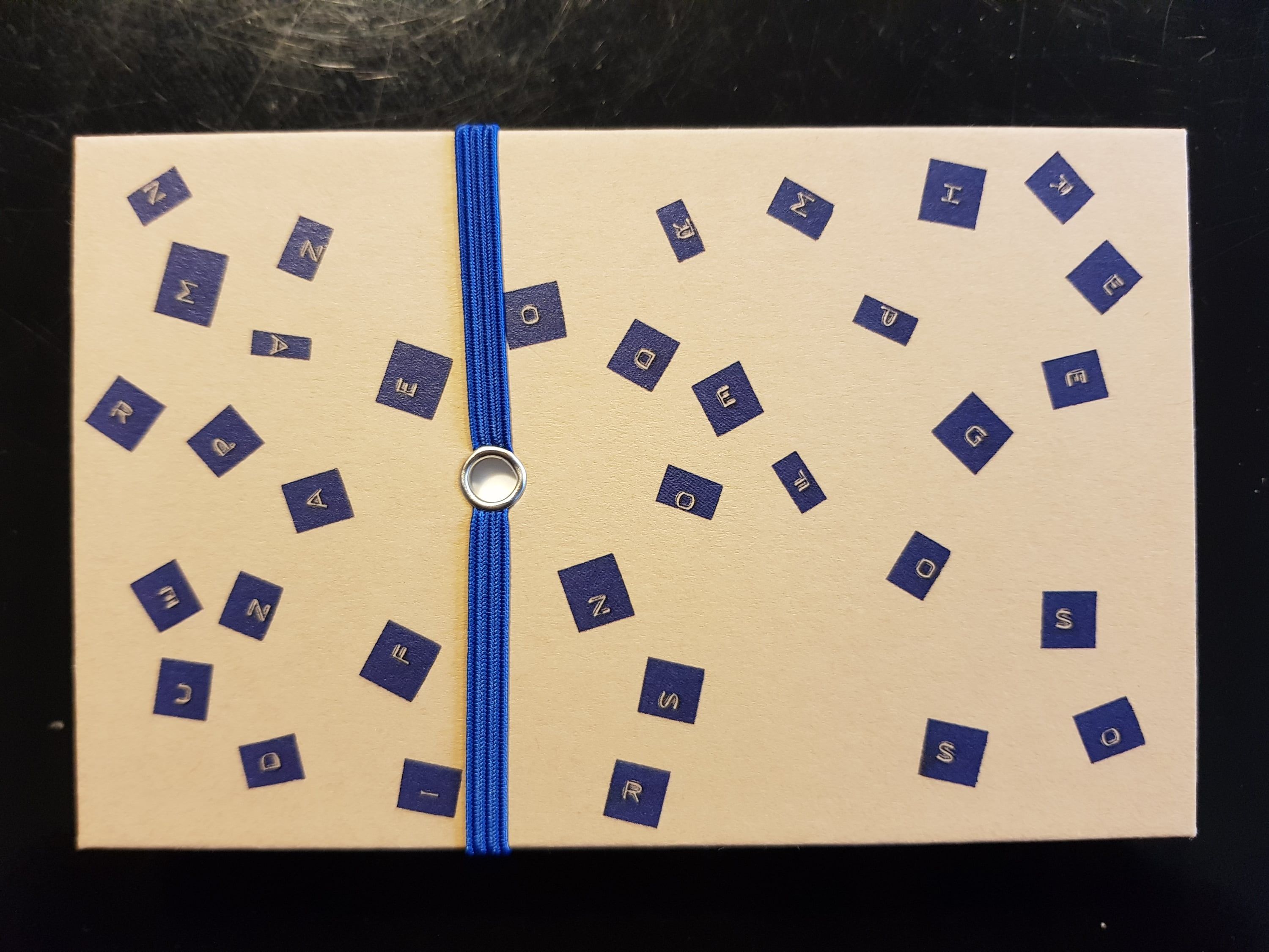 A second cover of the same booklet: the letters of the title are scattered in blue over the pale cover, and a blue elastic band with a metal eyelet runs over the middle.