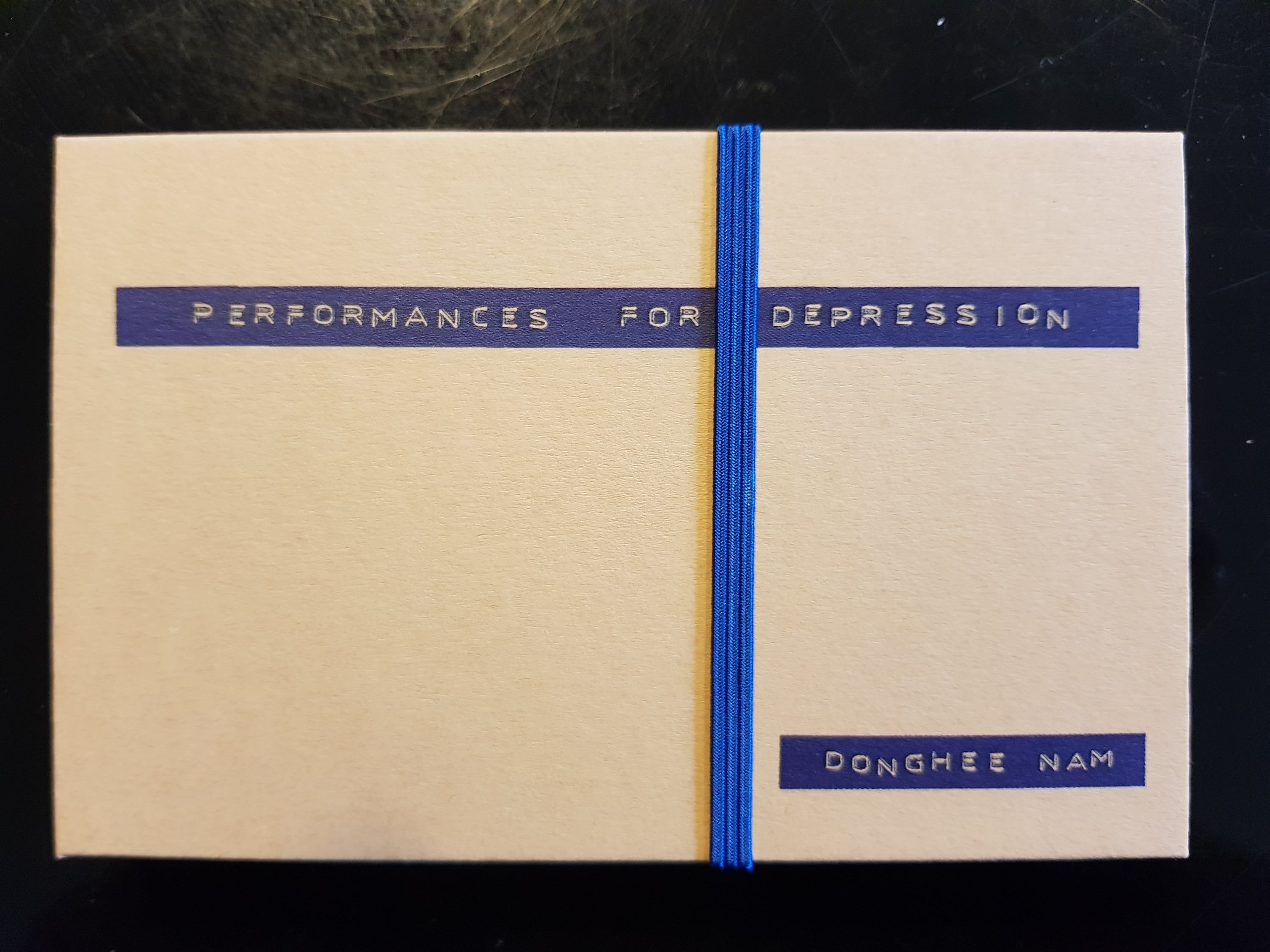 A small closed booklet with a pale cover on a dark table; blue label tape across it reads PERFORMANCES FOR DEPRESSION and, lower right, DONGHEE NAM; a blue elastic band runs over the middle.