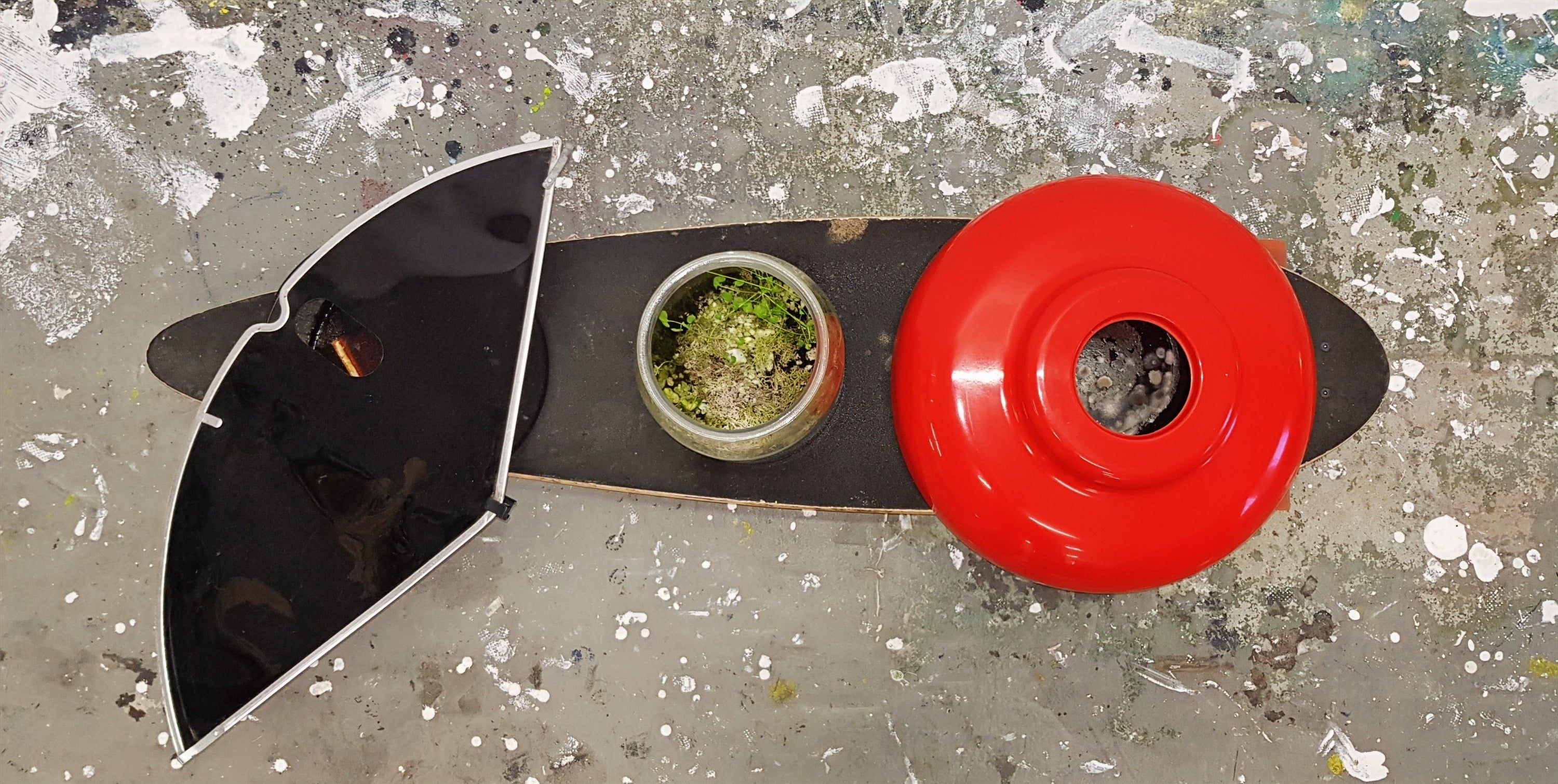 Das Longboard von oben auf einem farbbespritzten Boden: links eine dunkle Schale mit schrägem Deckel, in der Mitte ein Glas mit Pflanzen, rechts das rote Objekt mit einer dunklen Öffnung.