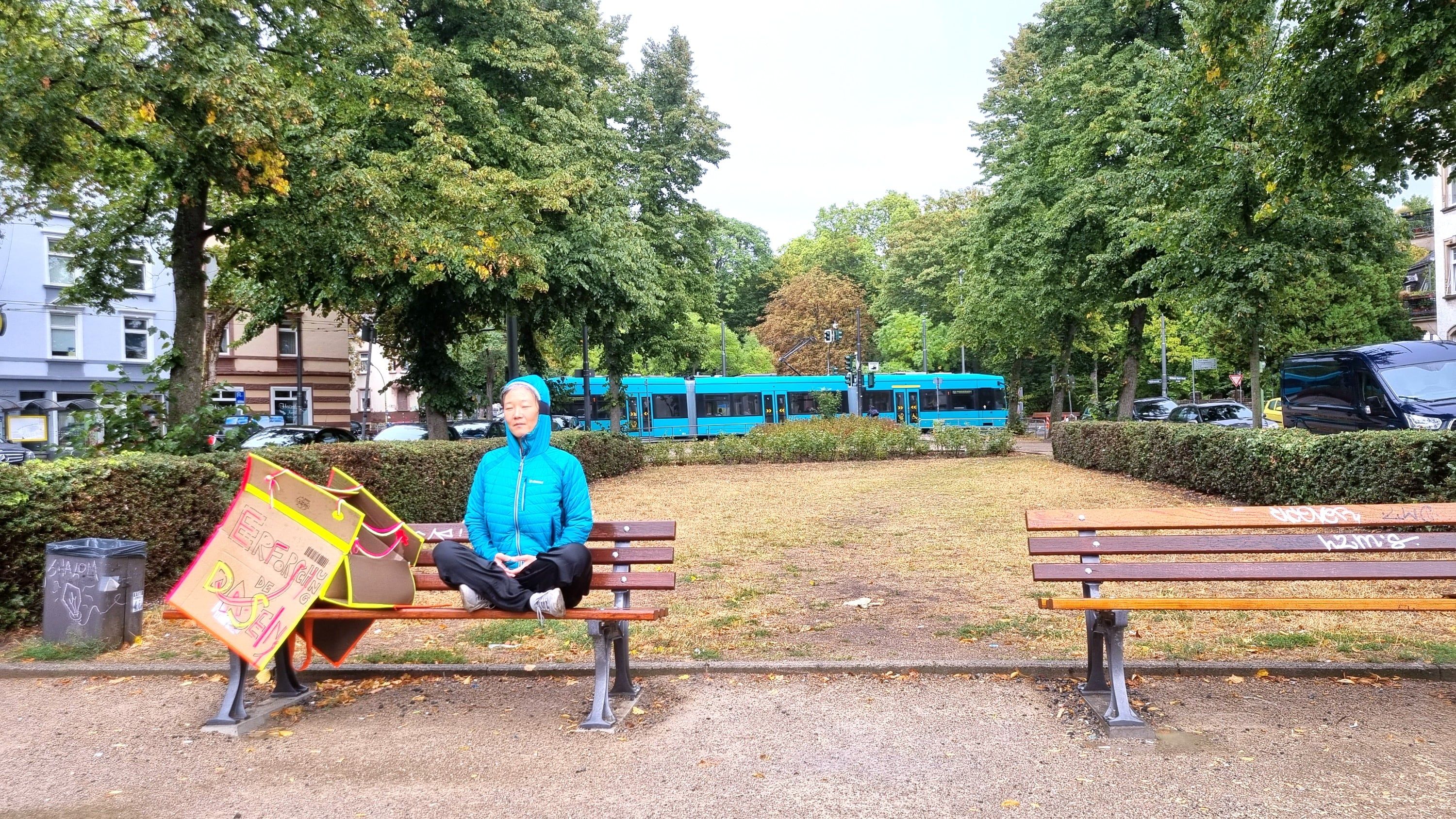 A person in a blue jacket sits on a park bench in an empty park, a yellow-orange sign leaning against the bench beside them; trees and a blue tram in the background.