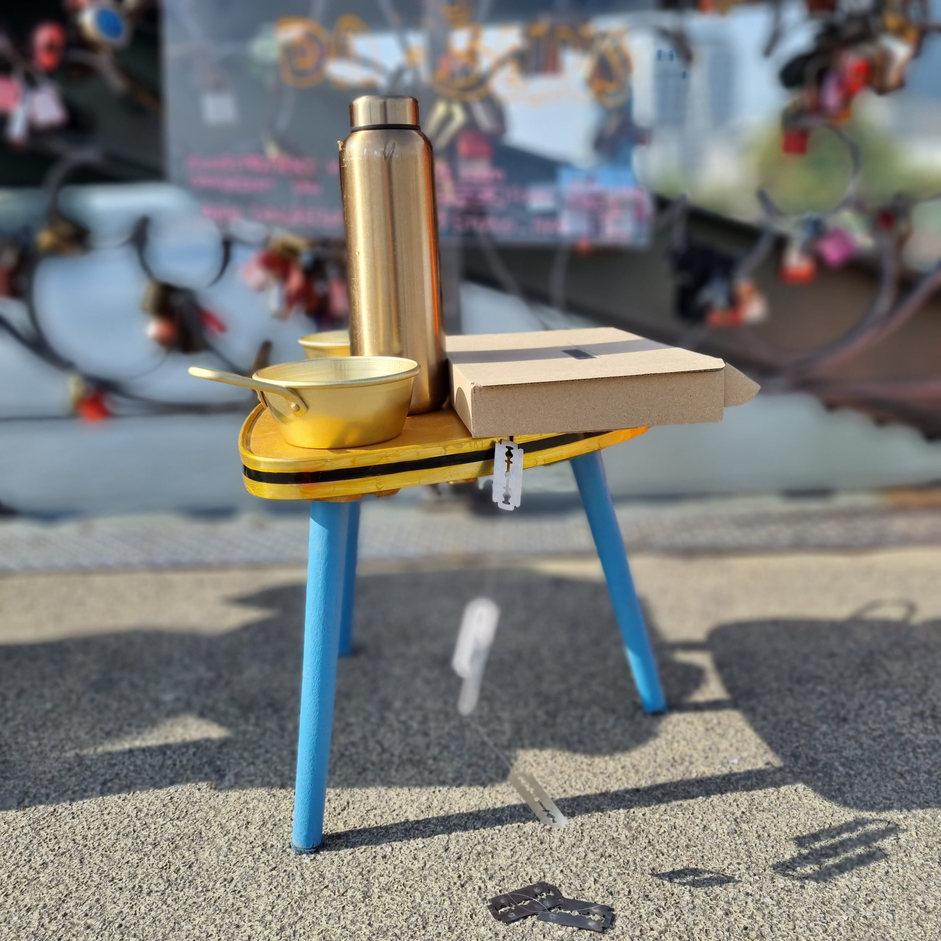 A small stool with blue legs and a golden top on a paved bridge, a golden bottle and a bowl with a wooden board standing on it; behind it the blurred love locks of the railing.