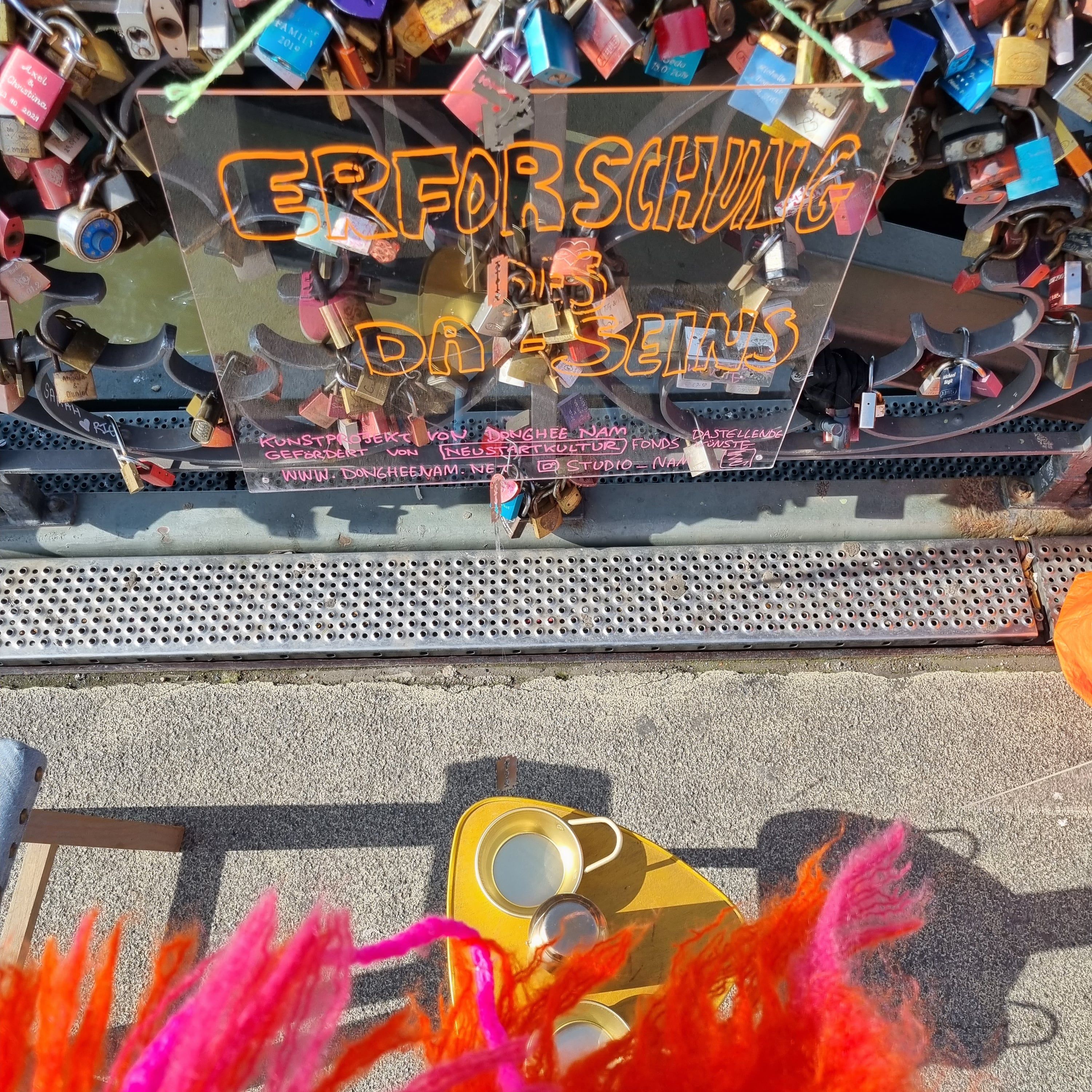 On the railing of a bridge covered in love locks stands a transparent sheet with orange lettering; in front of it a golden bowl and a golden bottle, below them pink feathers.