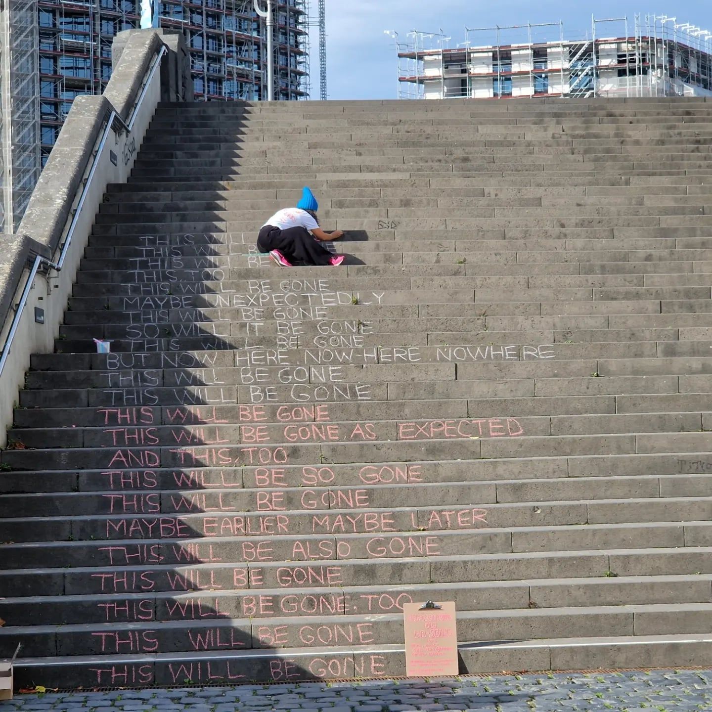 A wide concrete staircase seen from below, a person in a blue hood crouching near the top; faint chalk writing repeating the words THIS WILL BE GONE covers the steps.