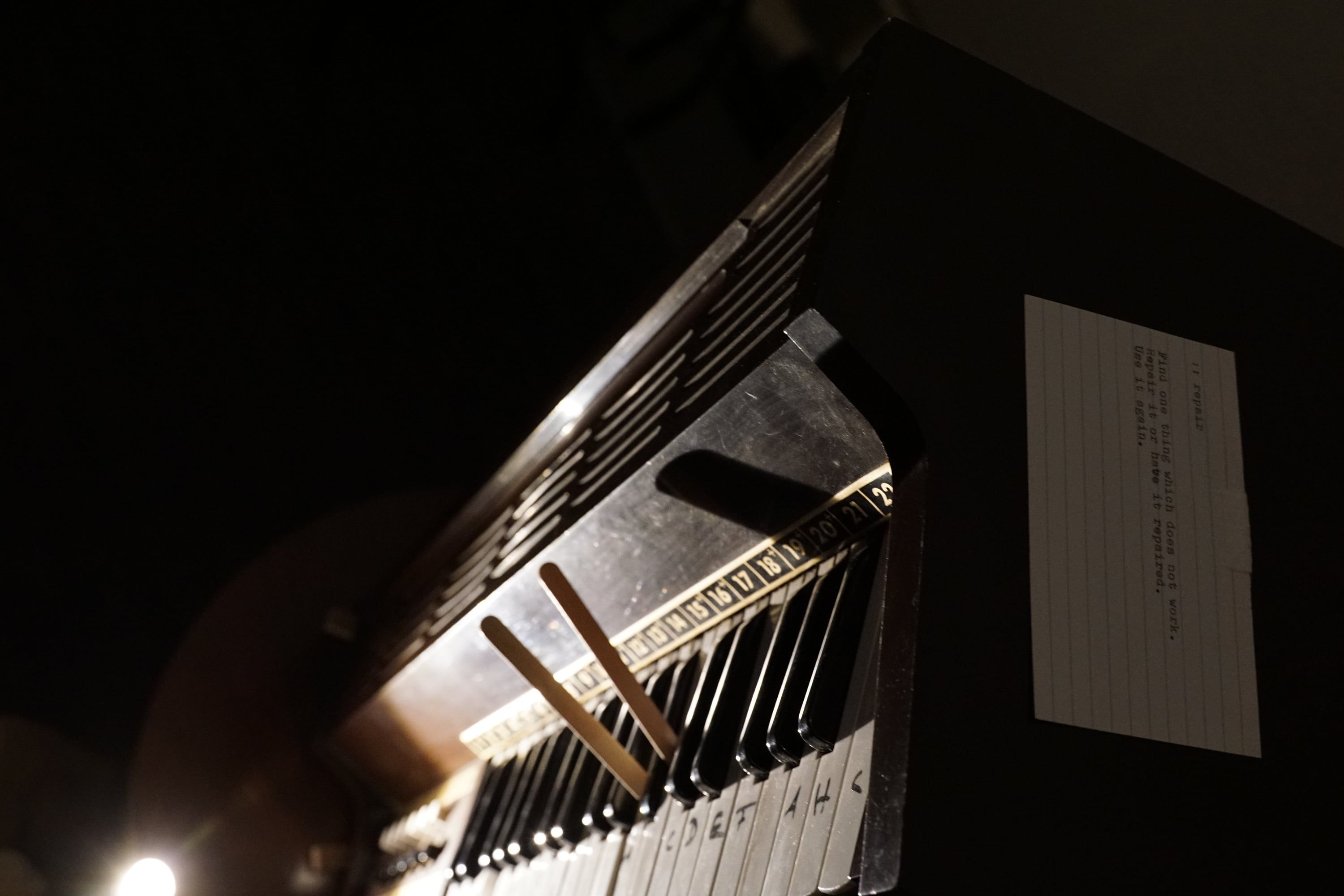 The keys of an old keyboard instrument in the dark, lit from one side, with an index card typed "repair" beside them