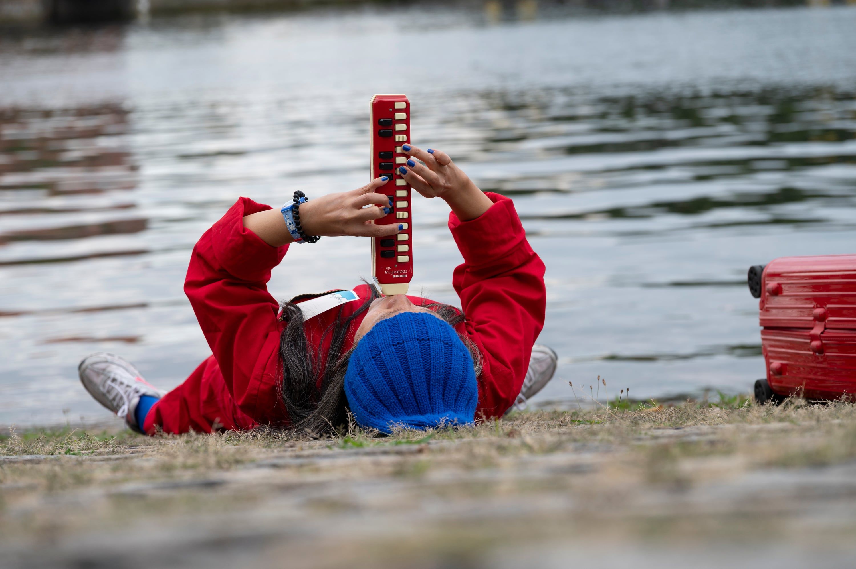 Donghee Nam lies on her back on a paved lakeside in a red overall and a blue knitted cap, playing a red melodica held upright above her face, a red suitcase at the right edge and grey water behind her