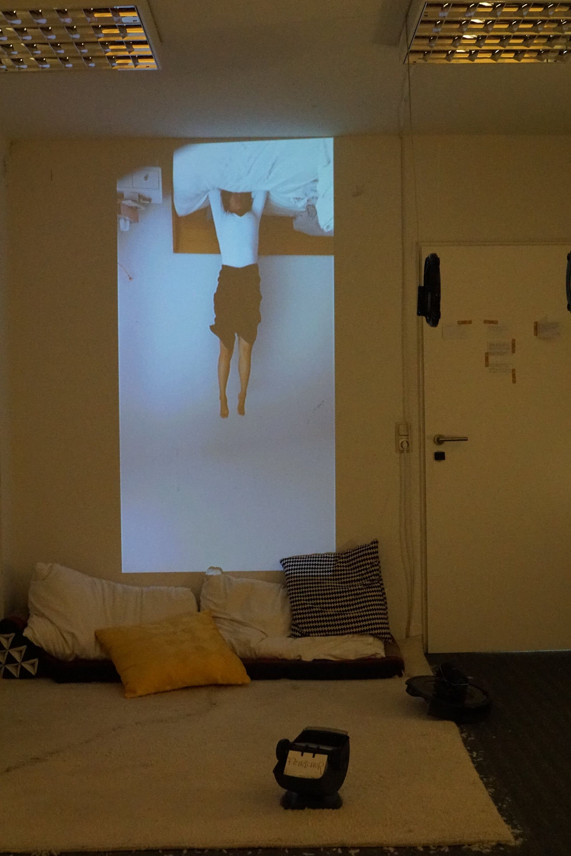 A room with cushions on a carpet; on the wall above them a projection of a person seen upside down, and in front, on the carpet, a rotary card file