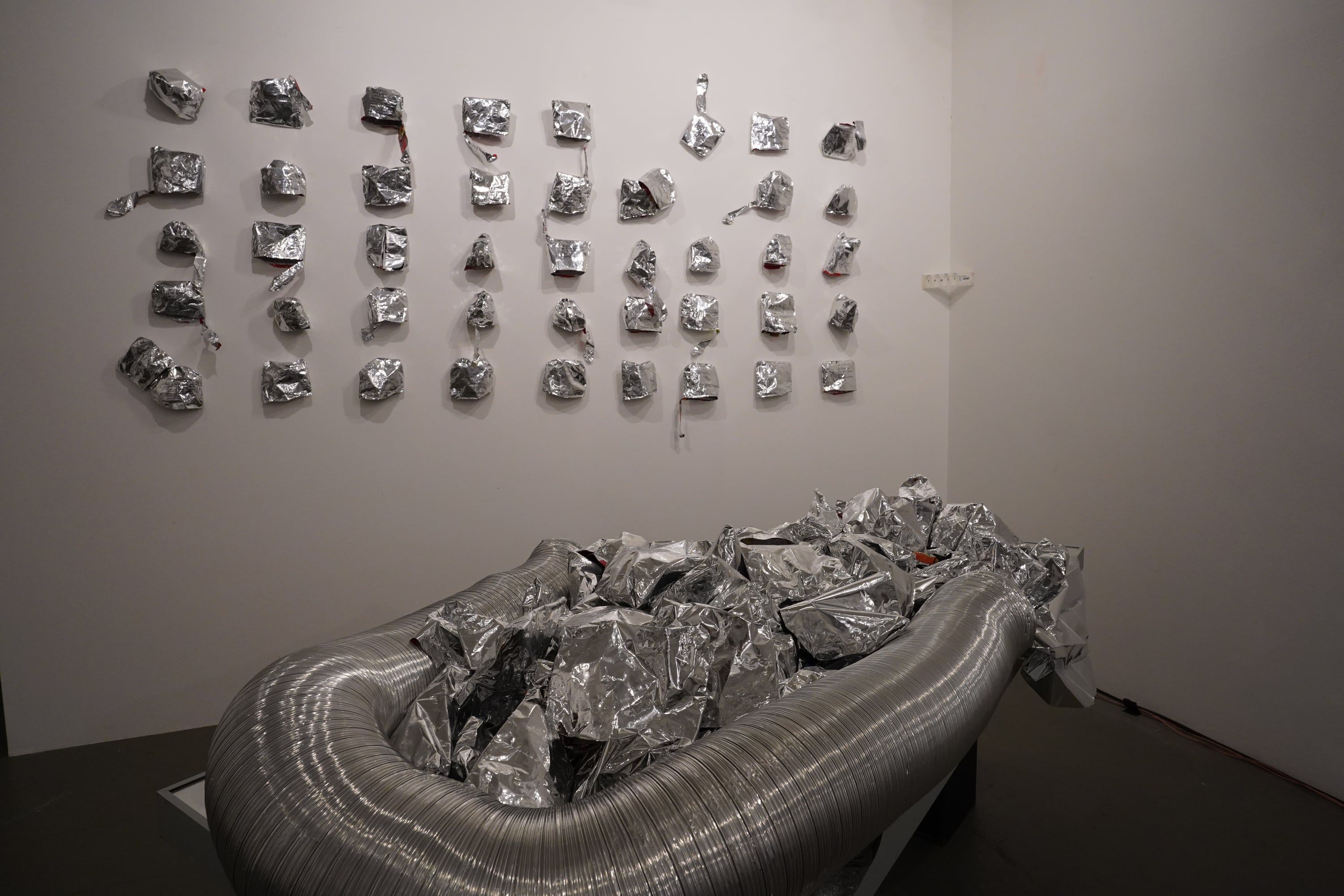 The room seen from the left: rows of crumpled aluminium pieces on a white wall, below them on a low table the silver tube filled with foil.