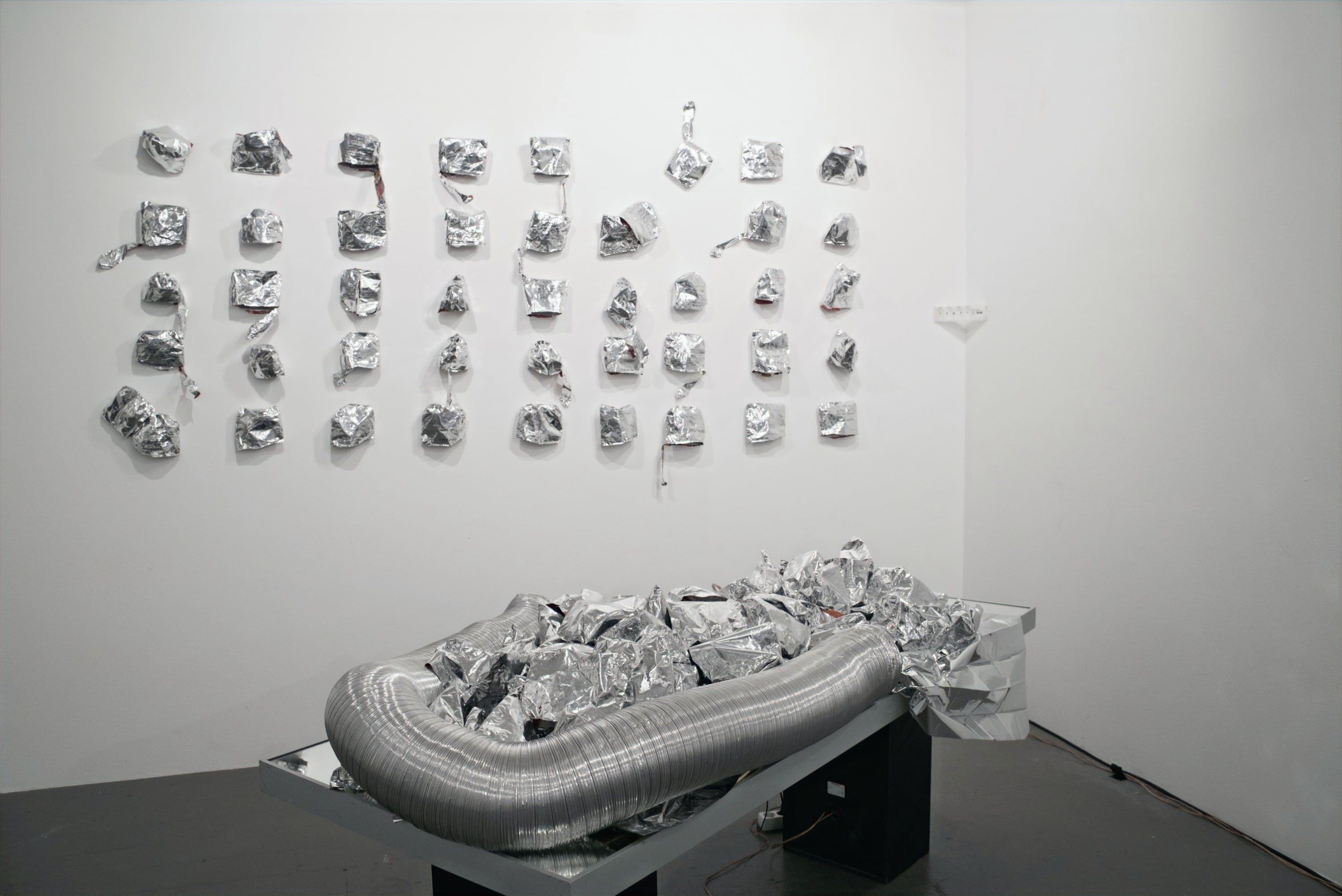 A corner of a white exhibition room: on the wall rows of crumpled silver bags, in front of them on a low table a ring of flexible aluminium duct filled with crumpled foil.