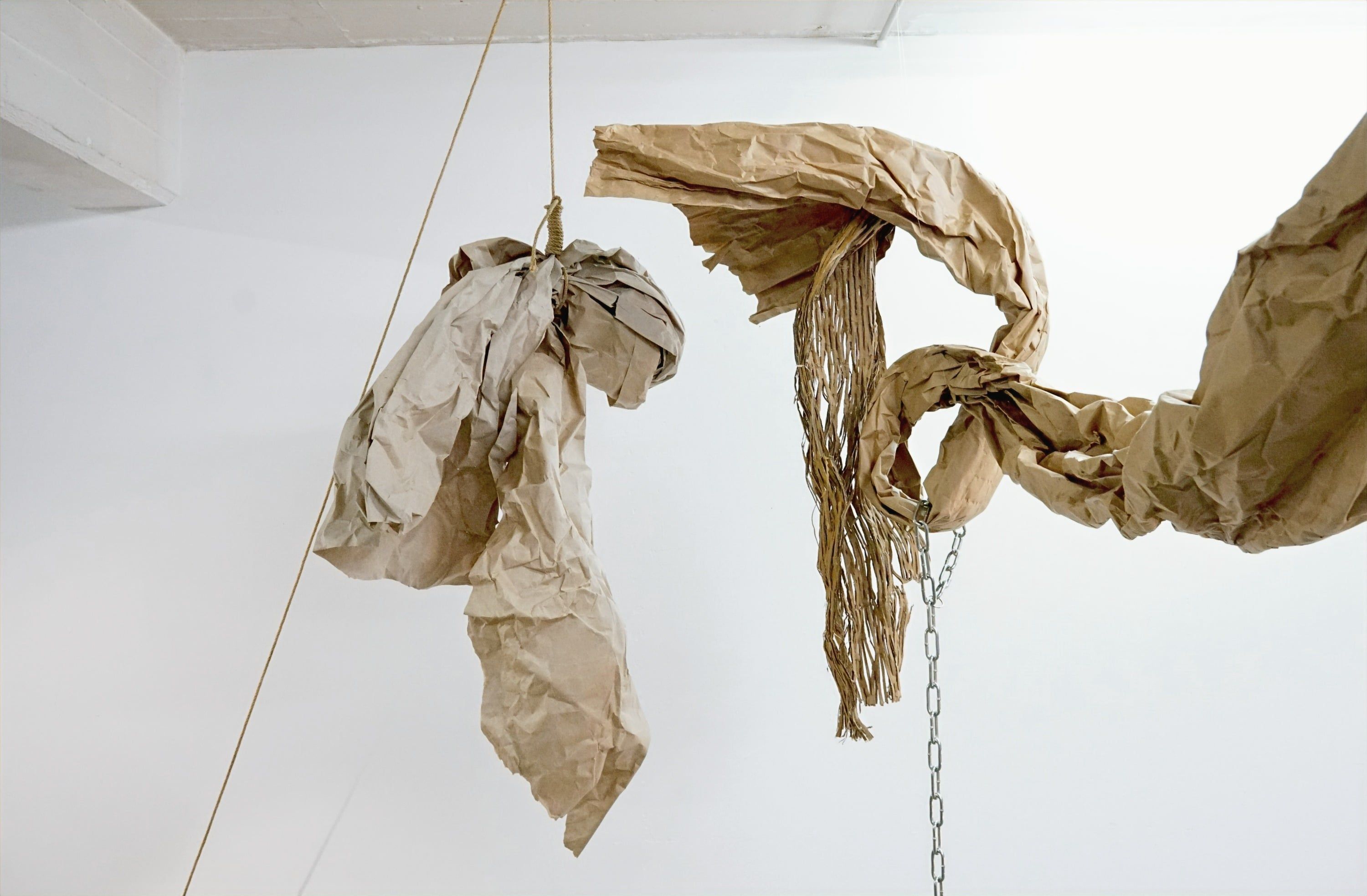 Ein Seil und eine Kette laufen von oben an einer weißen Wand entlang bis auf einen farbbespritzten Atelierboden, oben hängt Papier.