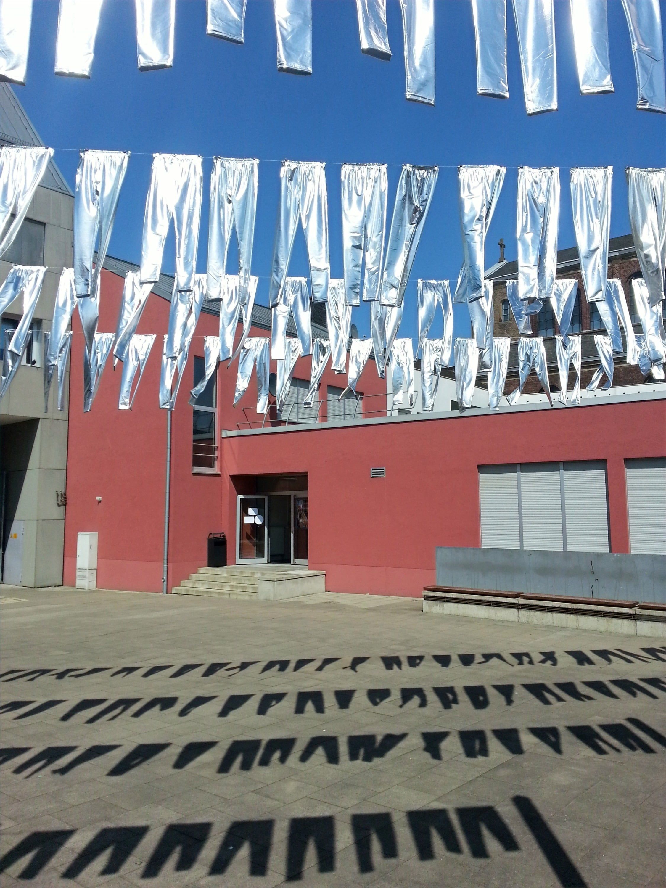 Reihen silberner Leggings hängen an Schnüren vor blauem Himmel über einem Hof mit rotem Gebäude; auf dem Pflaster darunter liegen ihre Schatten.