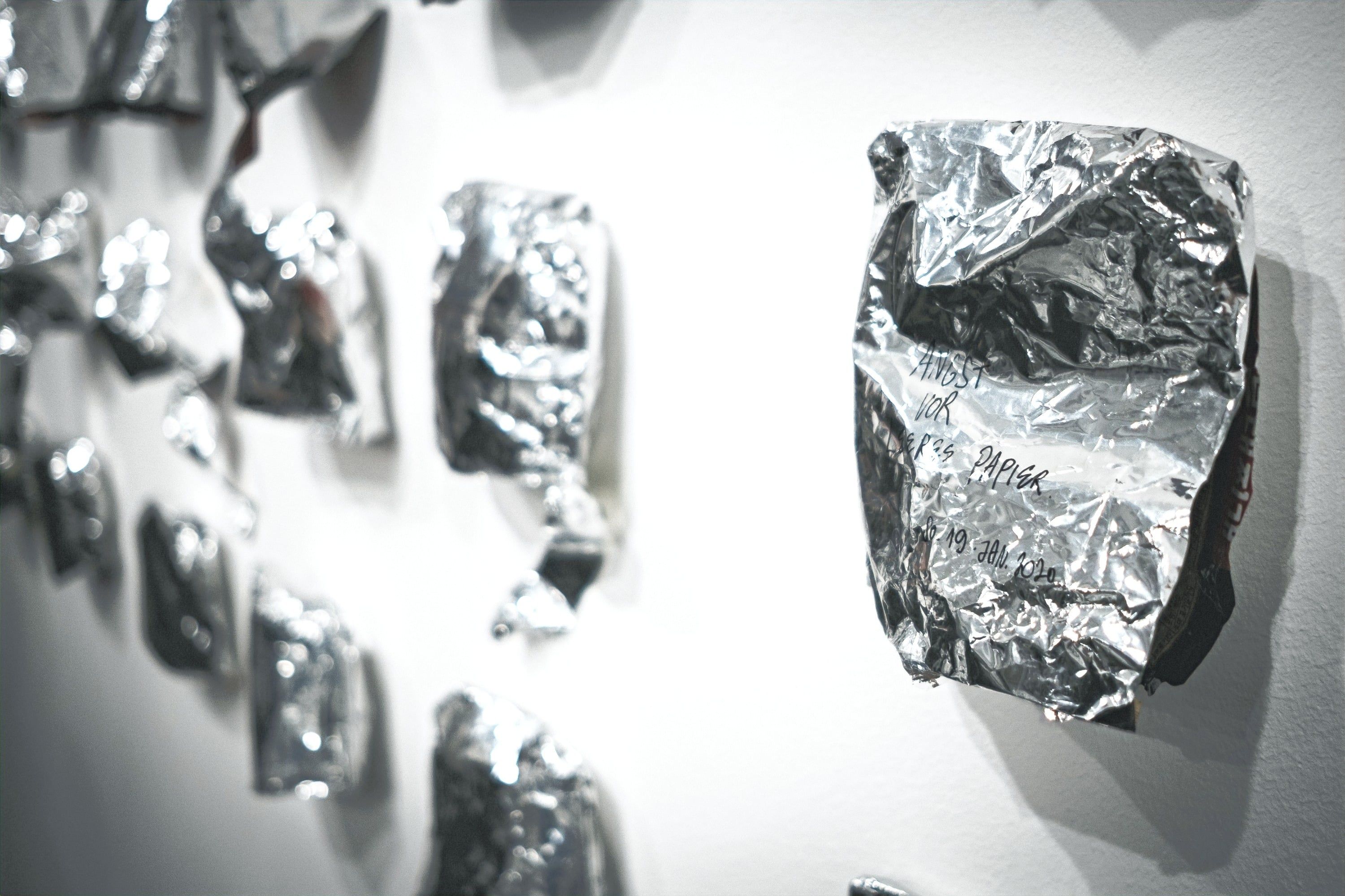 A written-on silver bag on a white wall seen from the side, more bags blurred in the foreground at left.