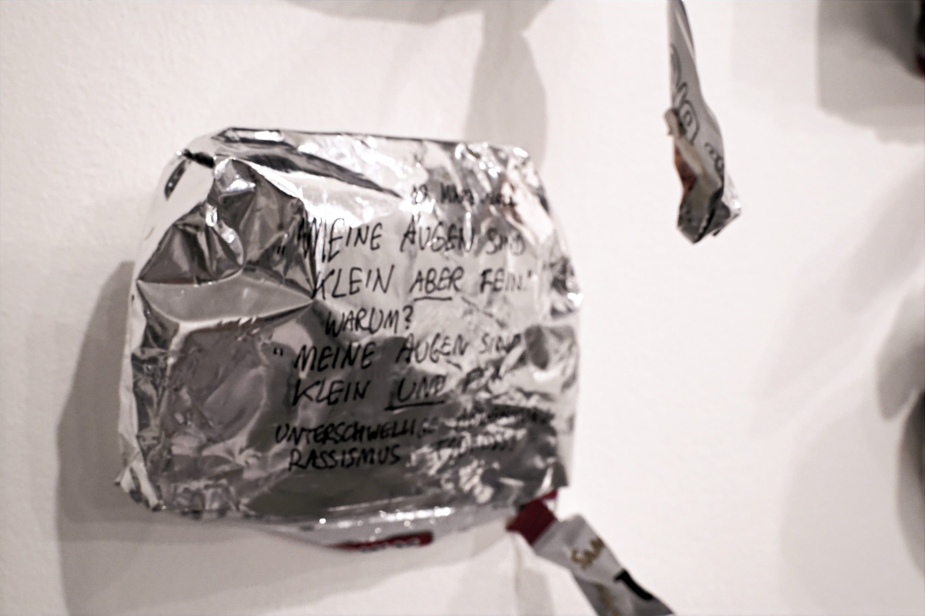 A crumpled silver bag on a white wall, written on in black marker; the German text is partly legible.