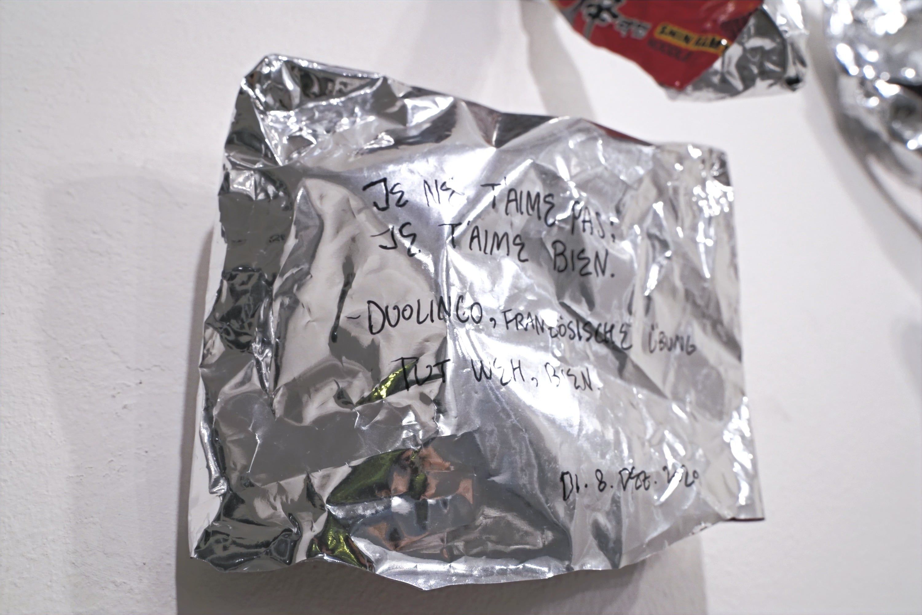 A crumpled silver bag on a white wall, densely covered with black marker writing in French and German; another bag with a red print reaches into the picture at the top.
