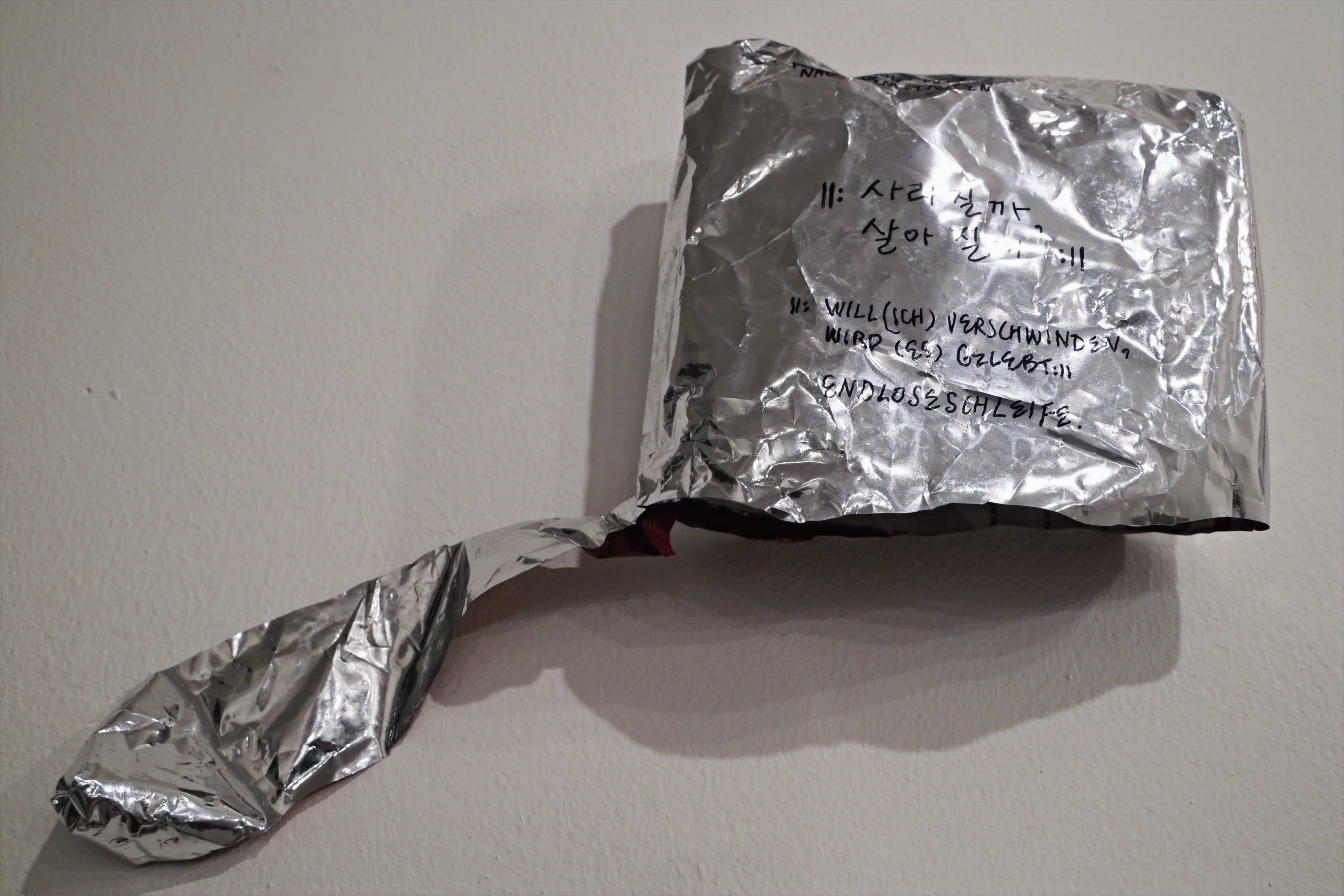 A crumpled silver bag on a wall, written on in black marker in Korean and German; a long strip of foil hangs from it down to the left.