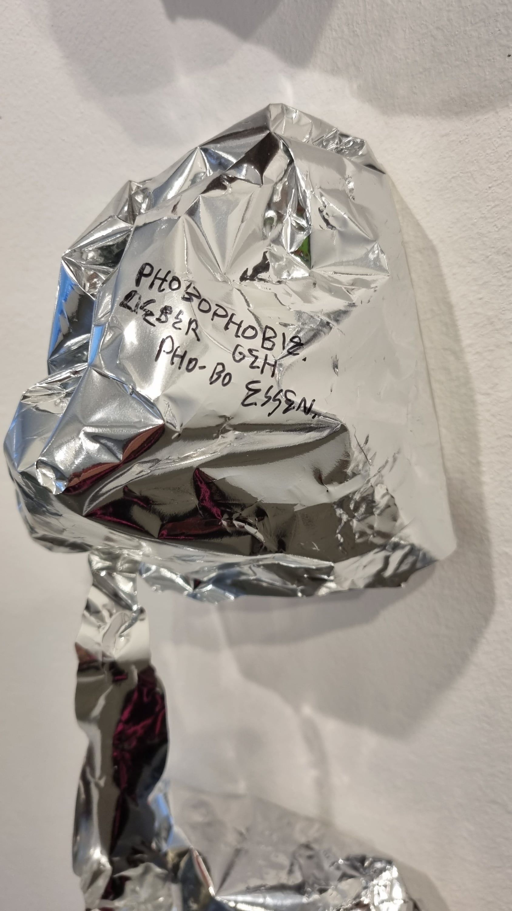 A crumpled silver bag on a white wall with a strip of foil hanging from it; the marker writing is partly legible.