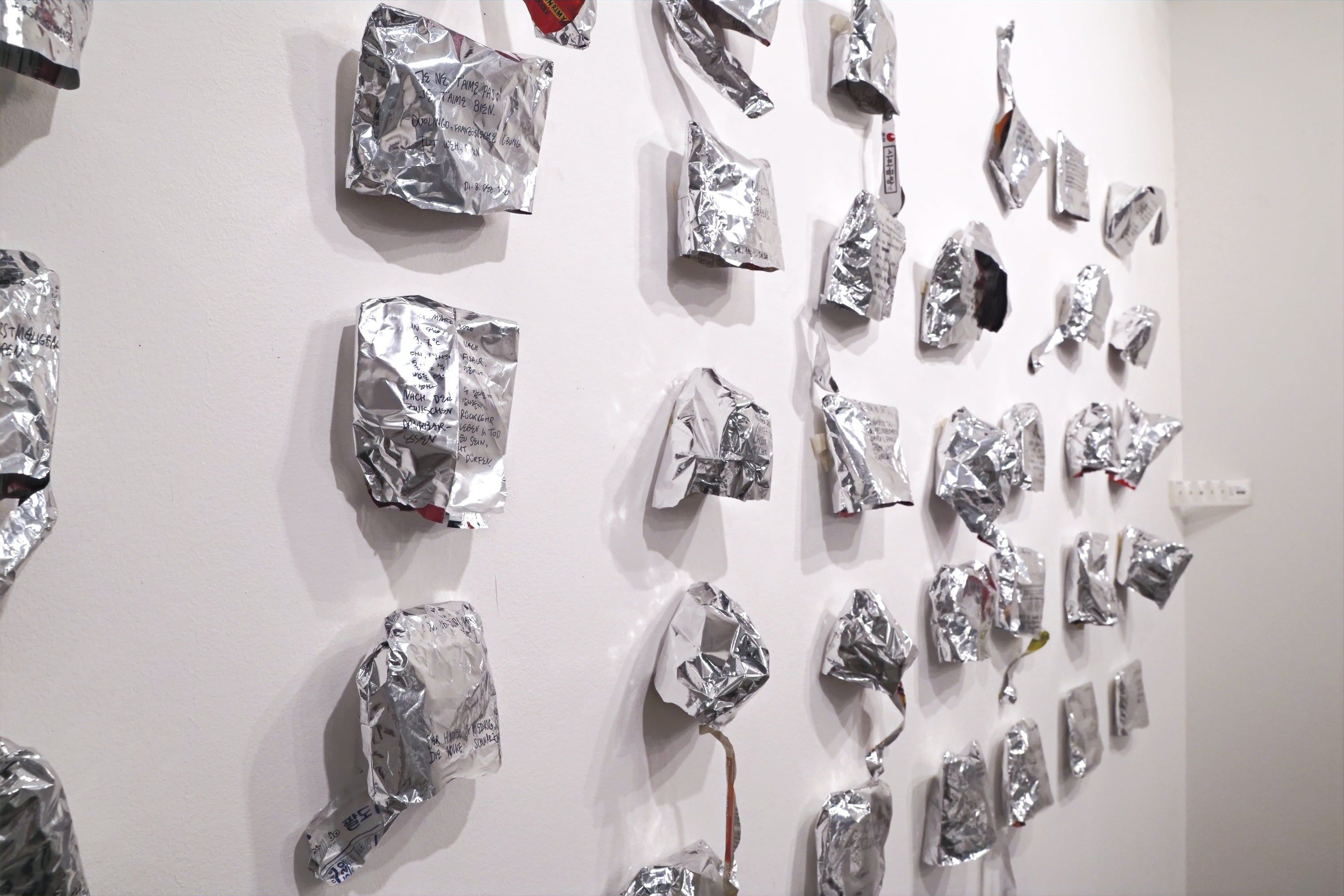 Seen at an angle from the side, crumpled silver noodle-soup bags hang in rows on a white wall.