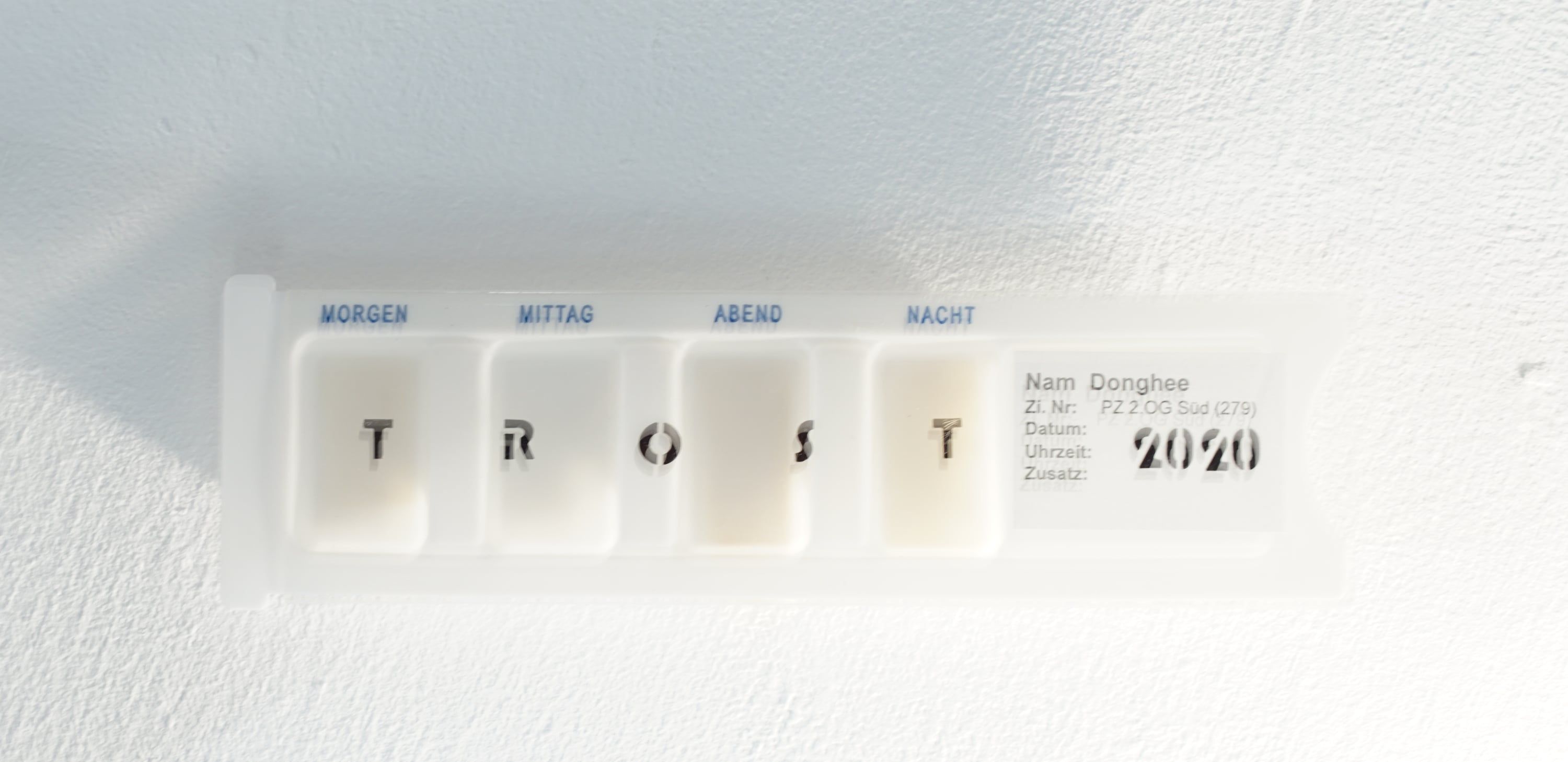 A white pill box on a wall with four compartments marked morning, noon, evening and night, holding the letters T R O S T, beside a label with the year 2020.