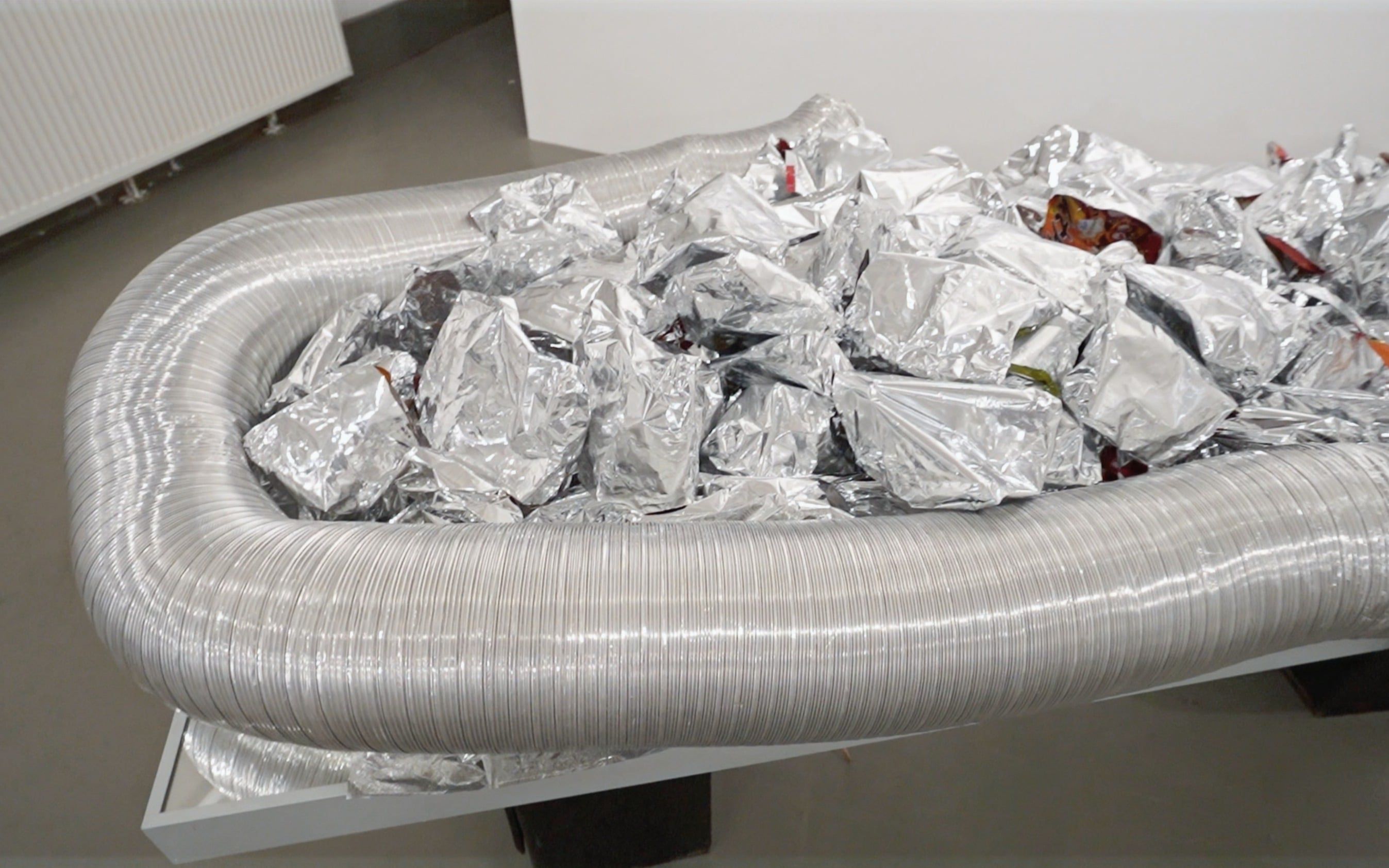A mirrored table seen from above, on it a ring of flexible aluminium duct holding a heap of crumpled silver bags.