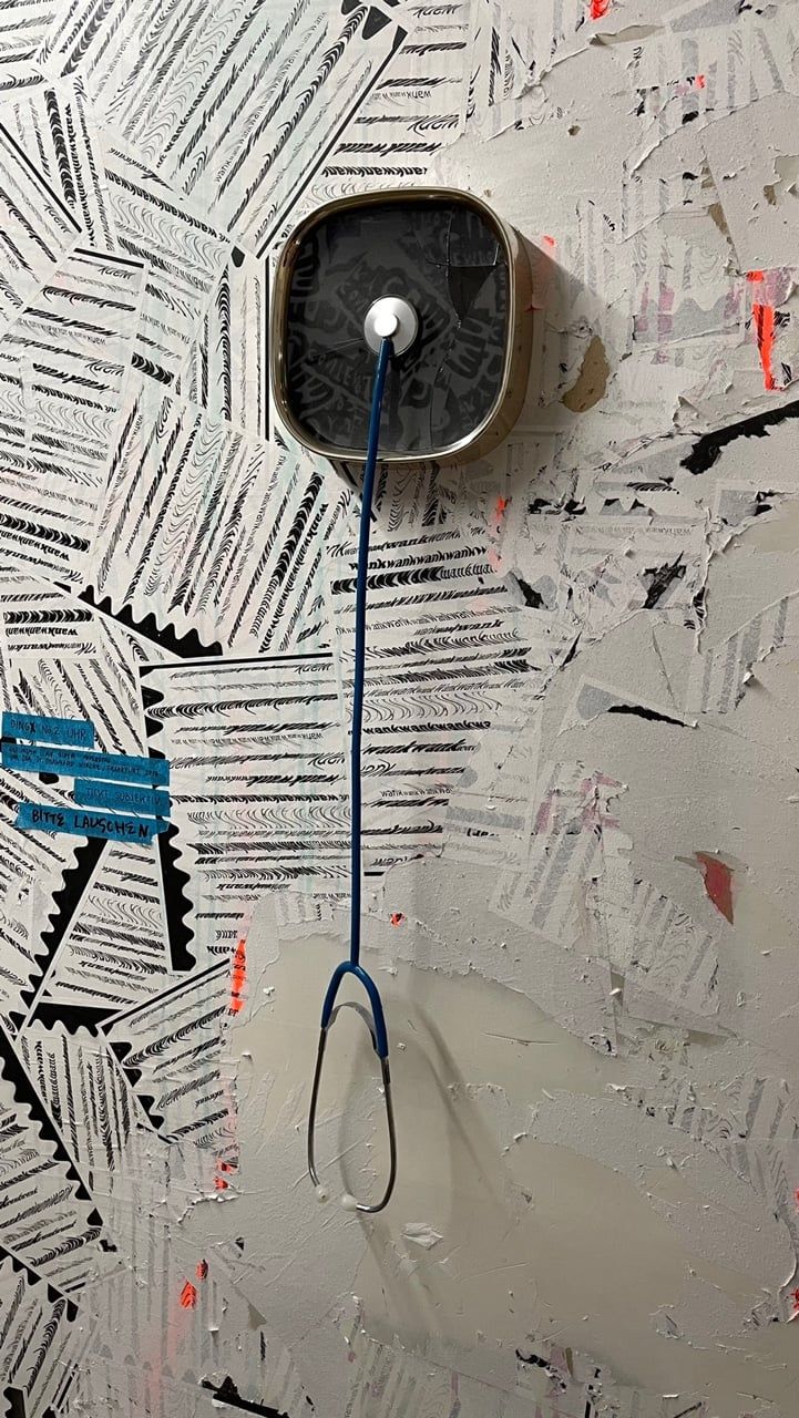 Dieselbe Uhr mit dem blauen Kabel an einer Wand, die von zerrissenen, schwarz bedruckten Papierstreifen bedeckt ist; links klebt ein Streifen blaues Klebeband mit Schrift.