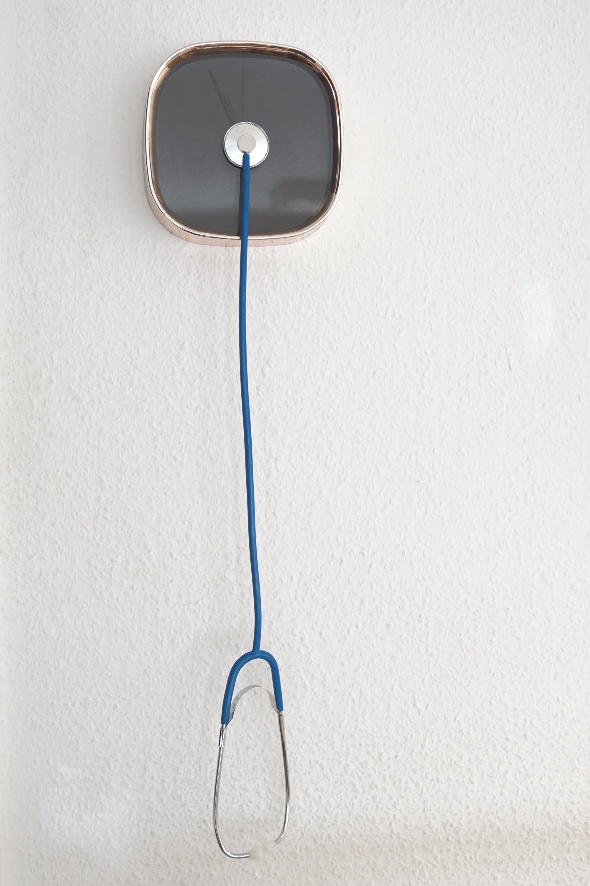 Eine Uhr mit abgerundetem, quadratischem Gehäuse an einer hellen Wand; aus ihrer Mitte läuft ein blaues Kabel nach unten und endet in einem metallenen Bügel.