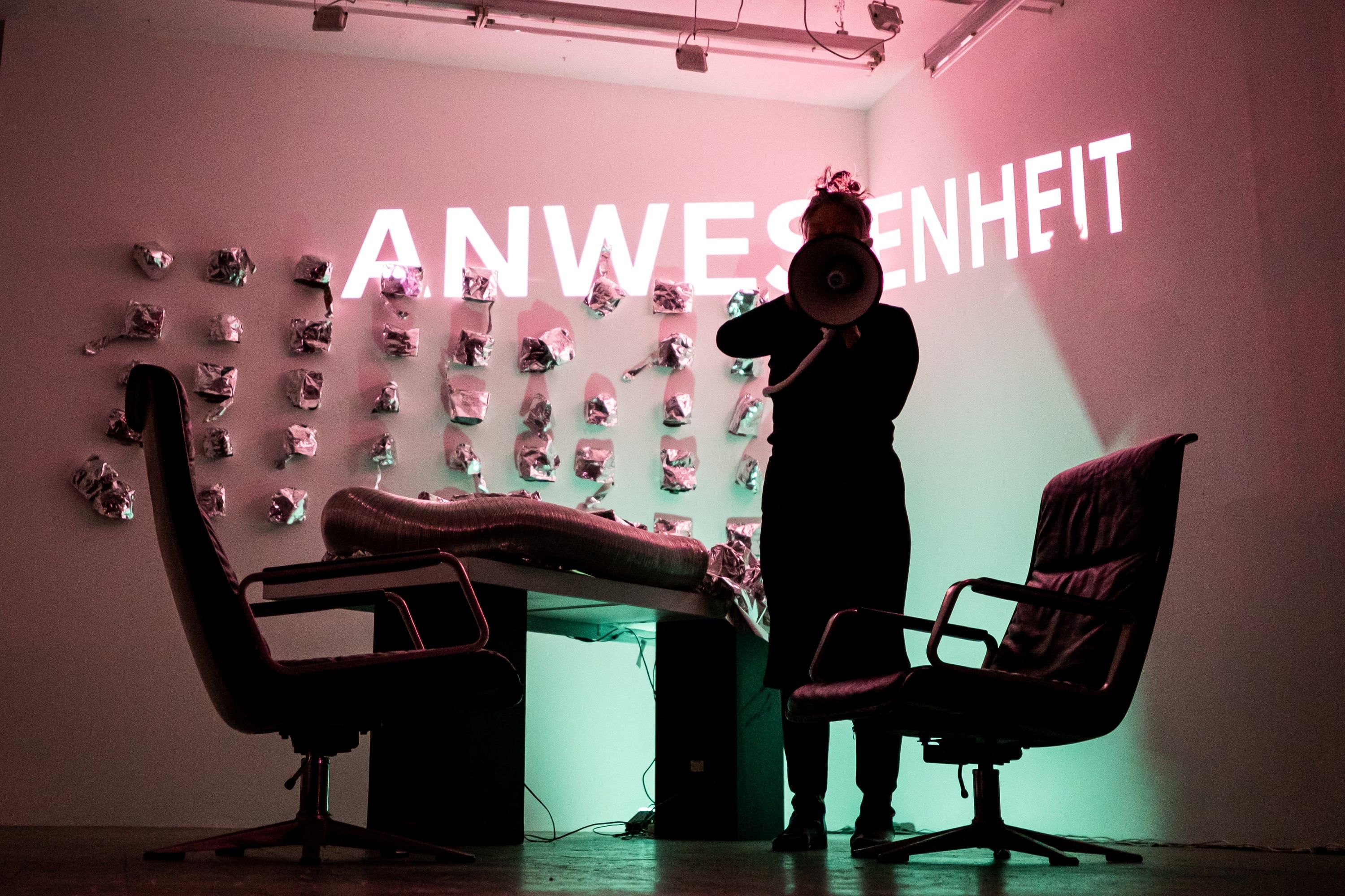 In pink and green light the person stands behind the megaphone between the two chairs, the word ANWESENHEIT projected across the wall of aluminium pieces.