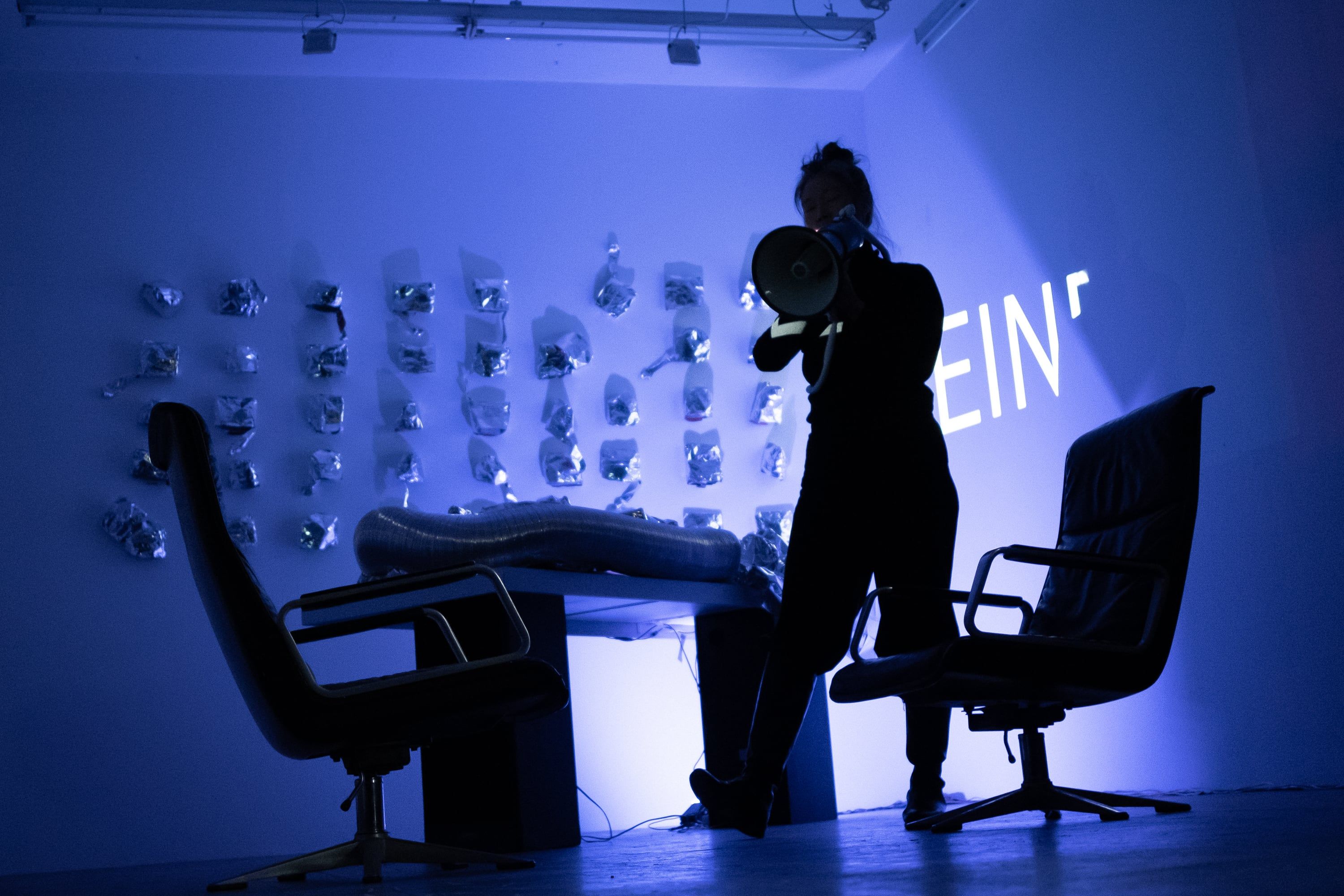 The same room in deep blue light: the person with the megaphone strides past the table, projected letters at the right on the wall.