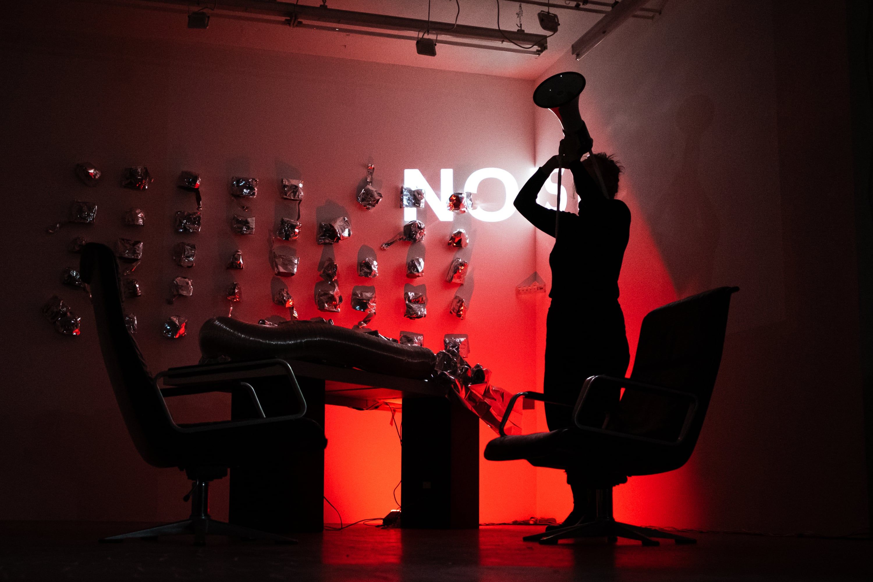 In red light the person holds the megaphone raised above the head, the projected letters NO on the wall behind.