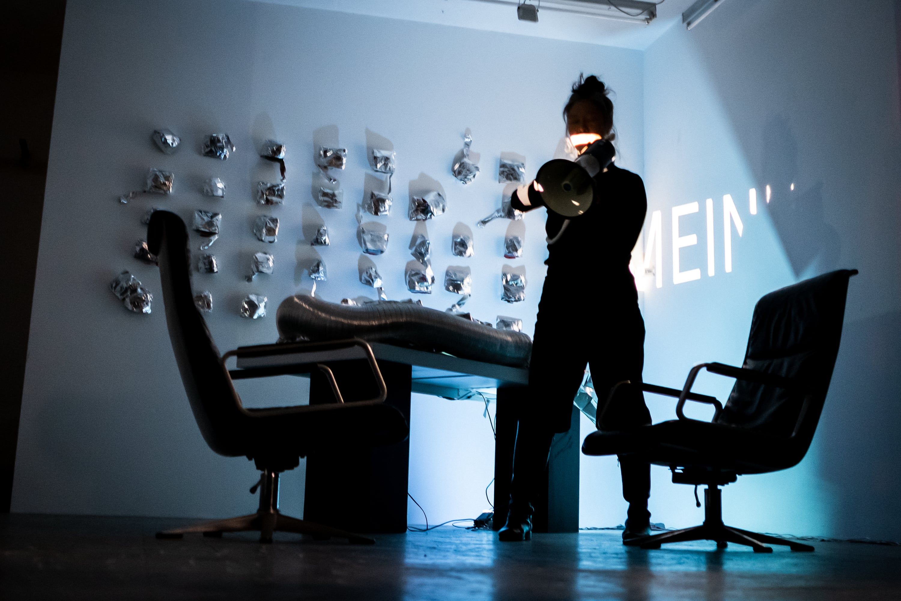 In pale blue light a person stands as a silhouette holding a megaphone to their face in front of a wall of hung aluminium pieces; an office chair on each side, between them a table with a ring of aluminium duct, projected letters partly legible on the wall.