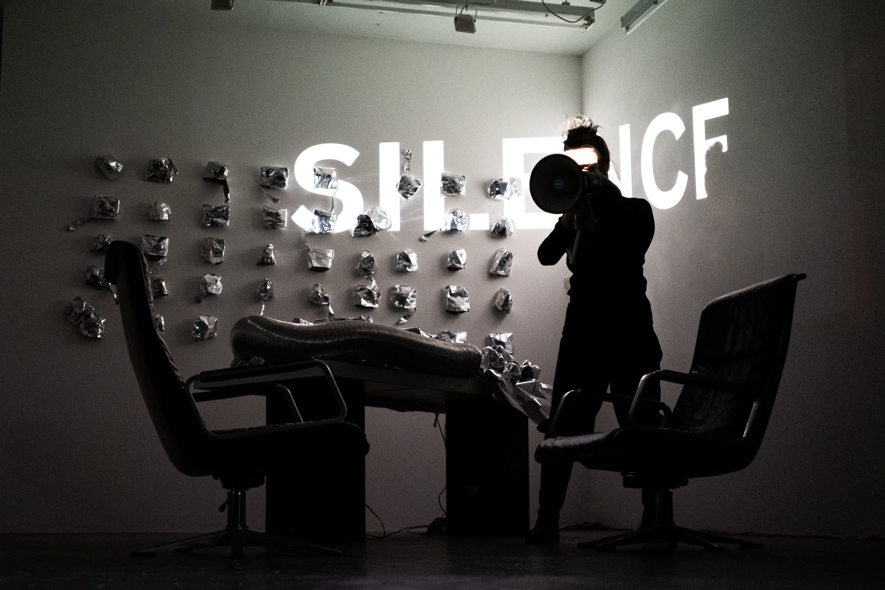 In grey light the person raises the megaphone, the projected word SILENCE partly hidden by the figure.
