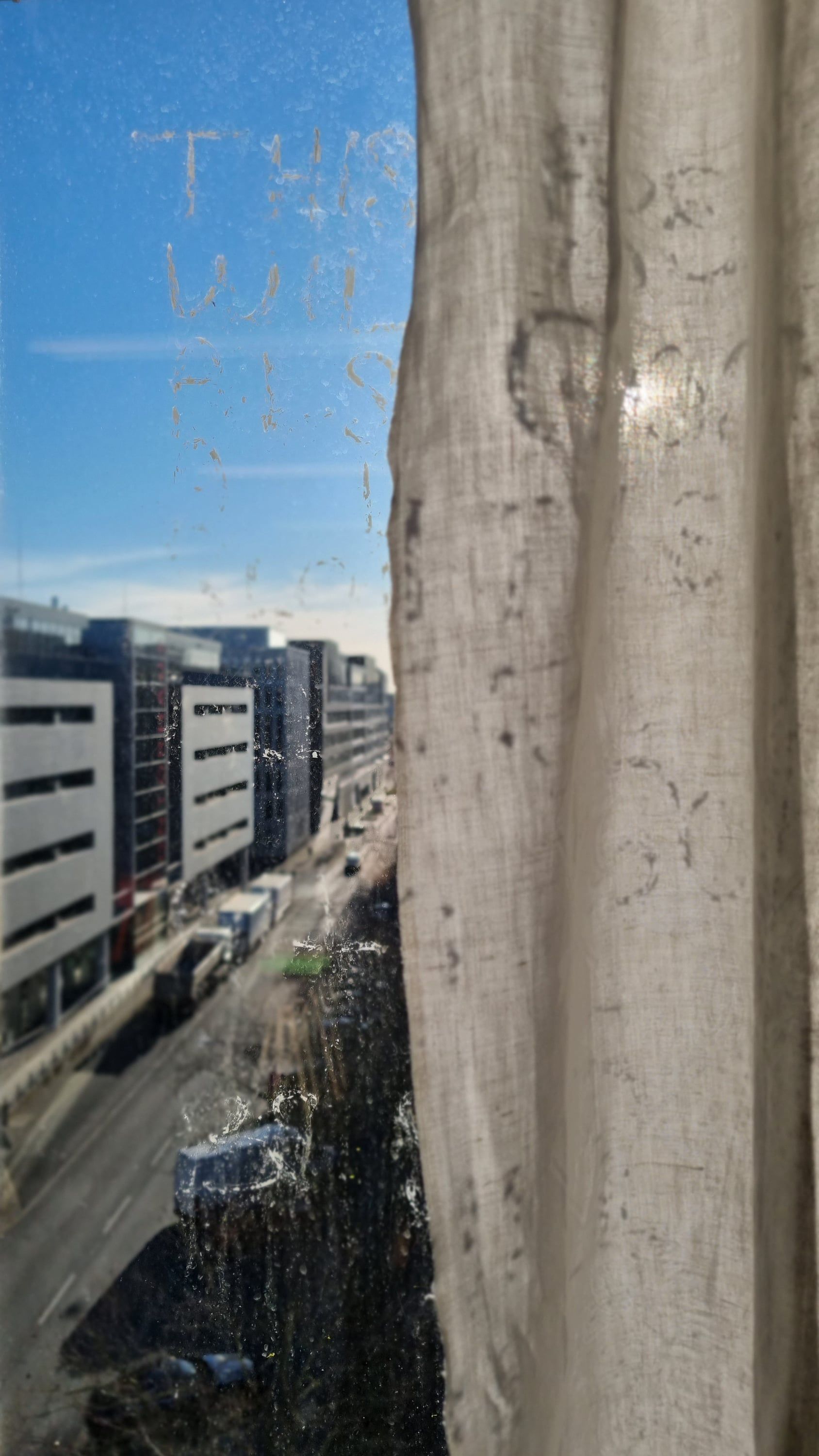 A window pane with letters in liquid chalk, only partly legible, looking down onto a street with a car park and trucks far below; a creased curtain hangs at the right.