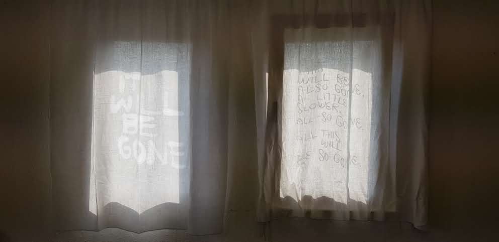 Two windows with sheer curtains, lit from behind in a dim room; on the left curtain the words IT WILL BE GONE in pale letters, on the right curtain several lines of writing.
